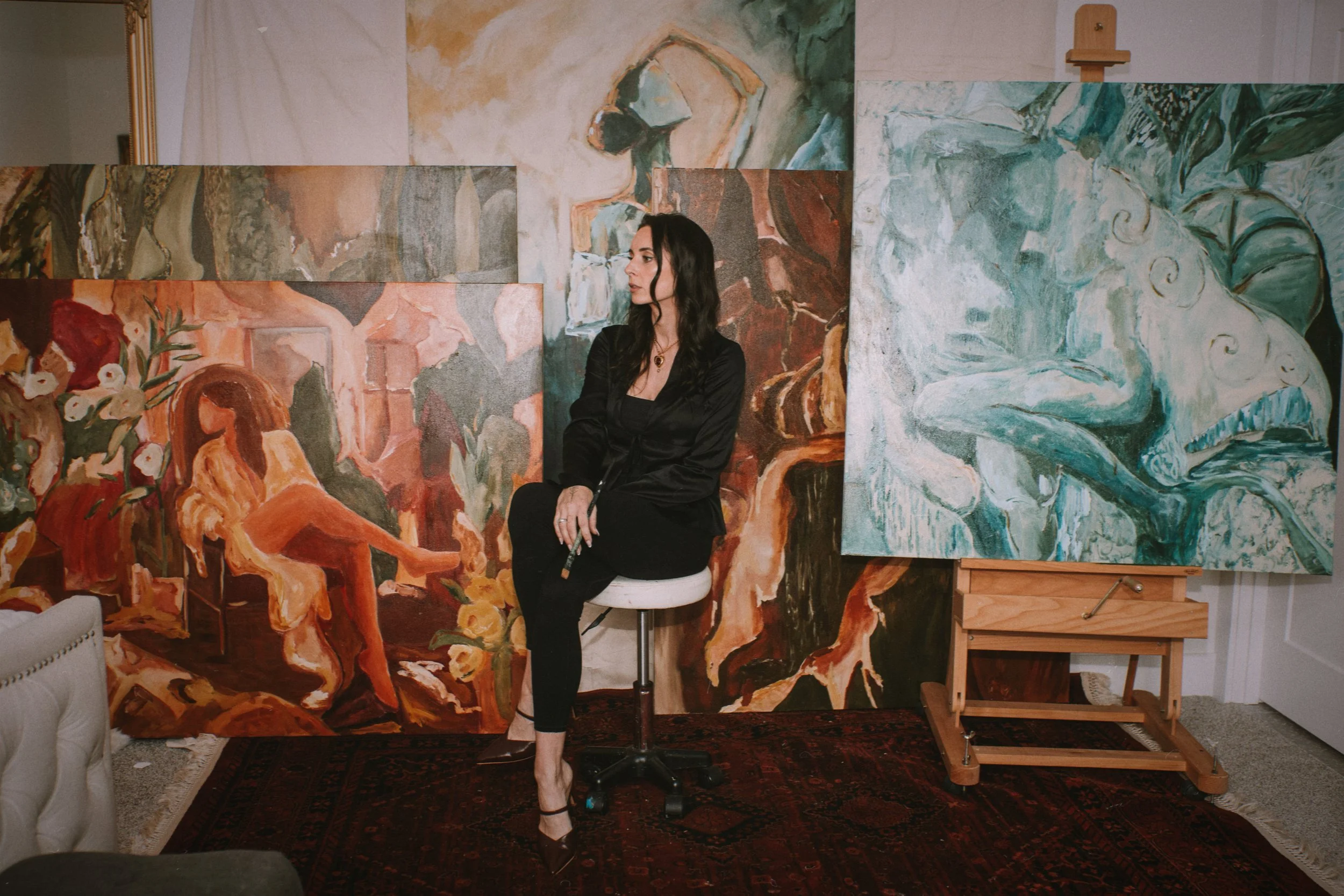 A woman with long dark hair sitting on an art studio stool in front of multiple large abstract paintings, wearing black clothing and high heels.