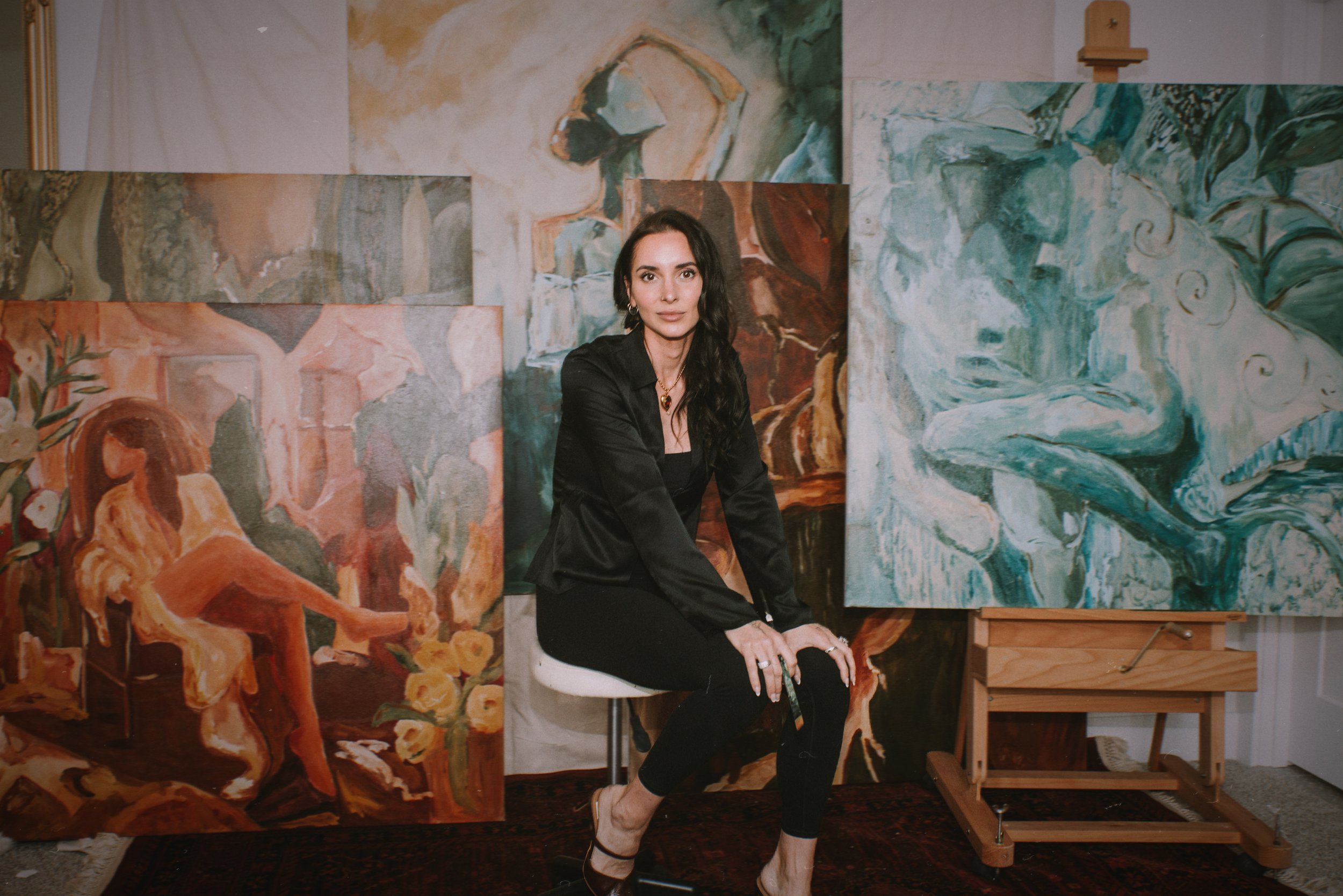 A local alaskan artist woman with dark hair and a black jacket sitting in front of multiple abstract paintings of figures and faces in an art studio.