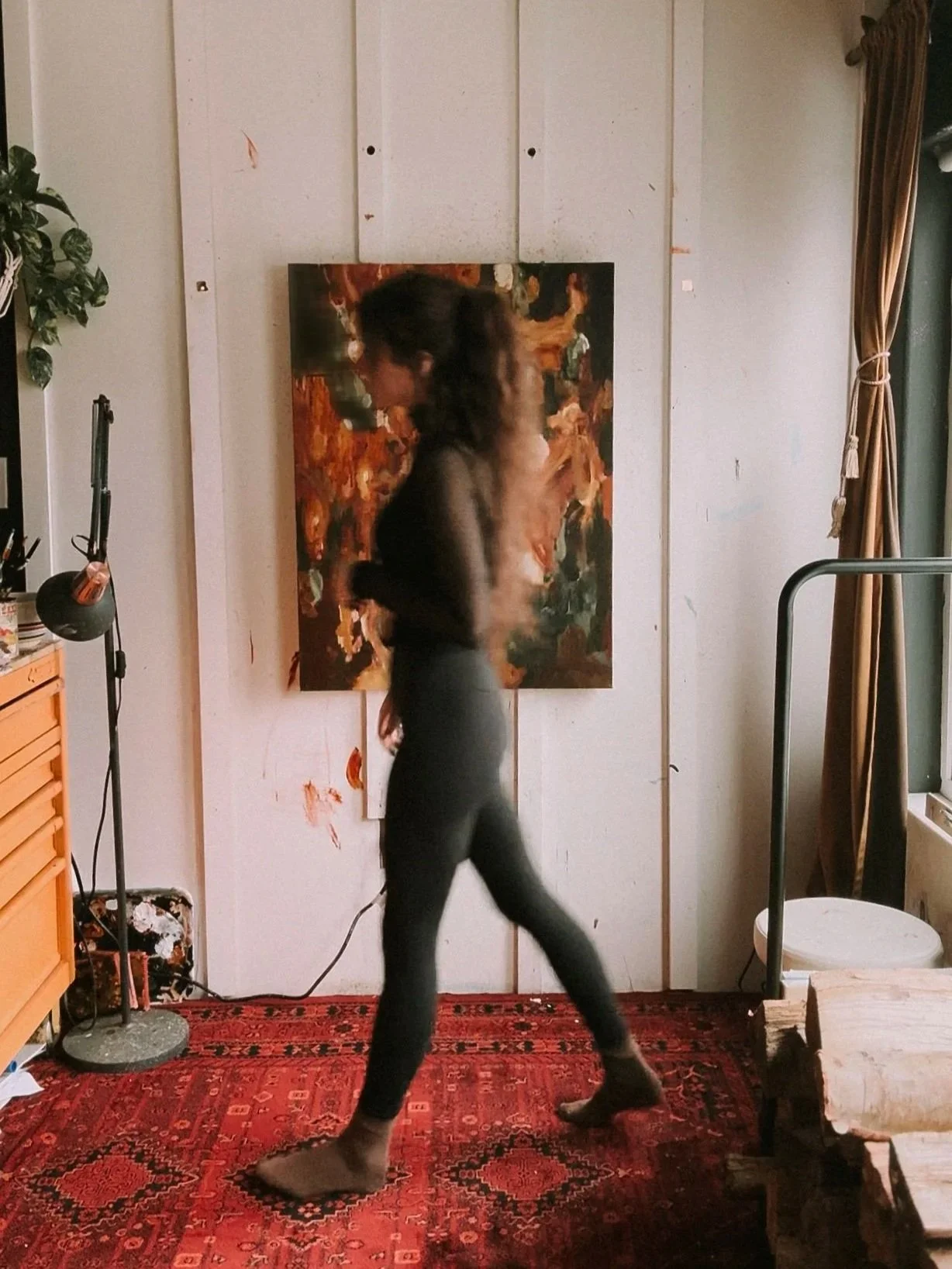 An artist walking barefoot in a studio with an abstract painting on the wall, a red patterned rug, and various art supplies and furniture.