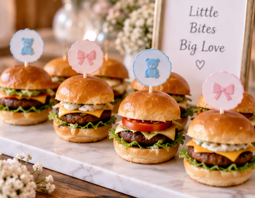 Mini sliders decorated with blue bear and pink bow toppers on a marble tray, with a framed sign reading "Little Bites Big Love" and floral decorations in the background.