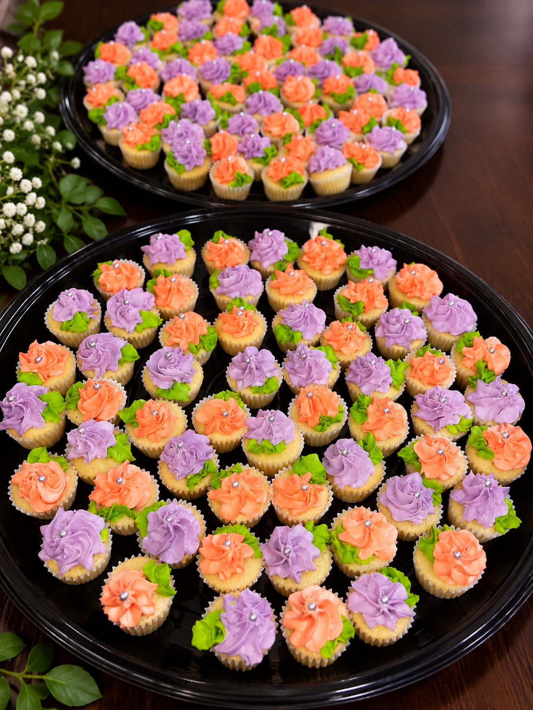 Tray of decorated cupcake treats with purple and orange frosting, green leaves, and silver edible beads, arranged on black trays on a wooden surface with green foliage nearby.