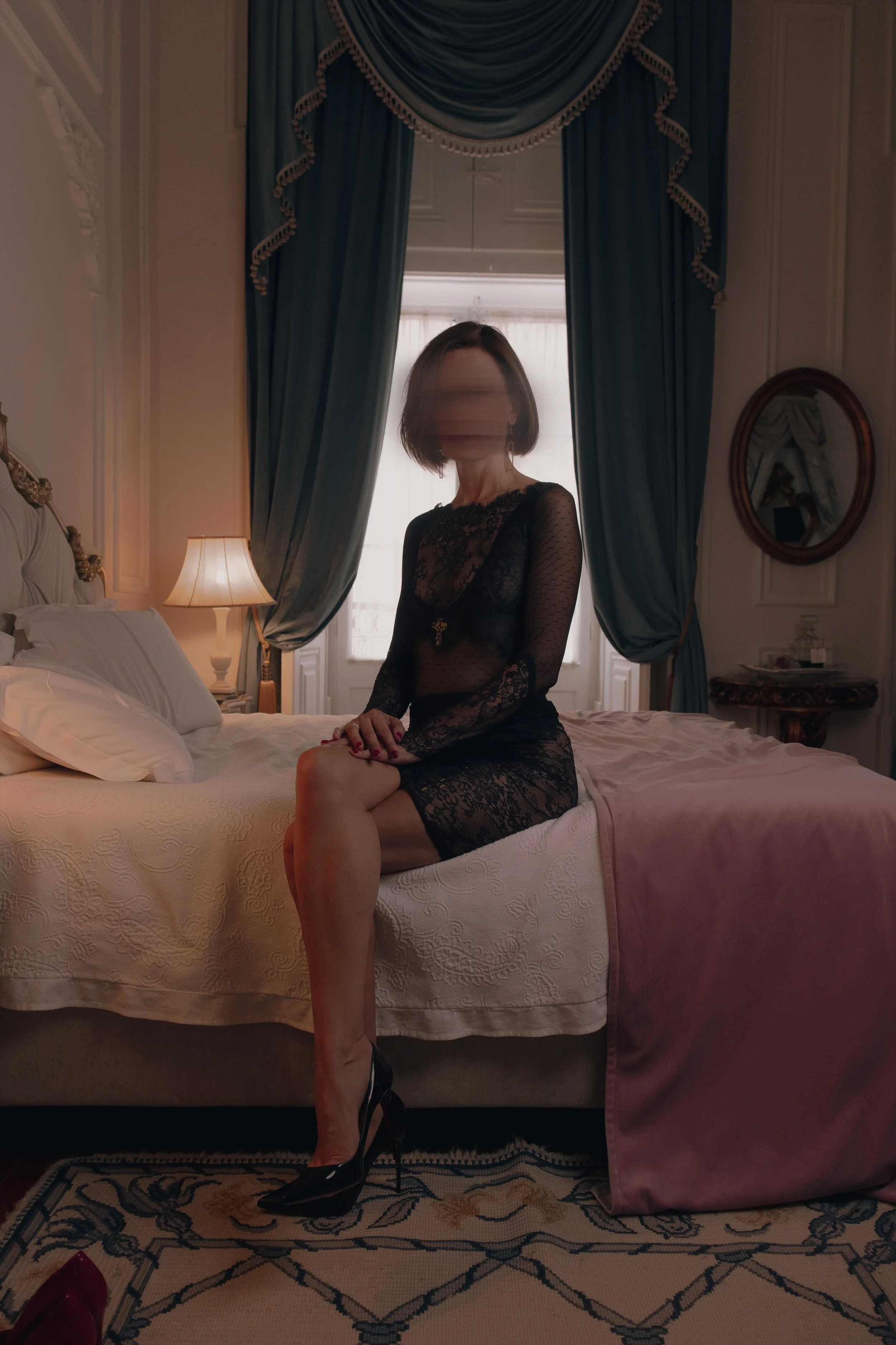 A Mistress sitting on the edge of a bed in a luxurious bedroom, wearing a black lace dress and black high heels, louboutins, with short brown hair, in front of a large window with blue curtains and a mirror on the wall.
