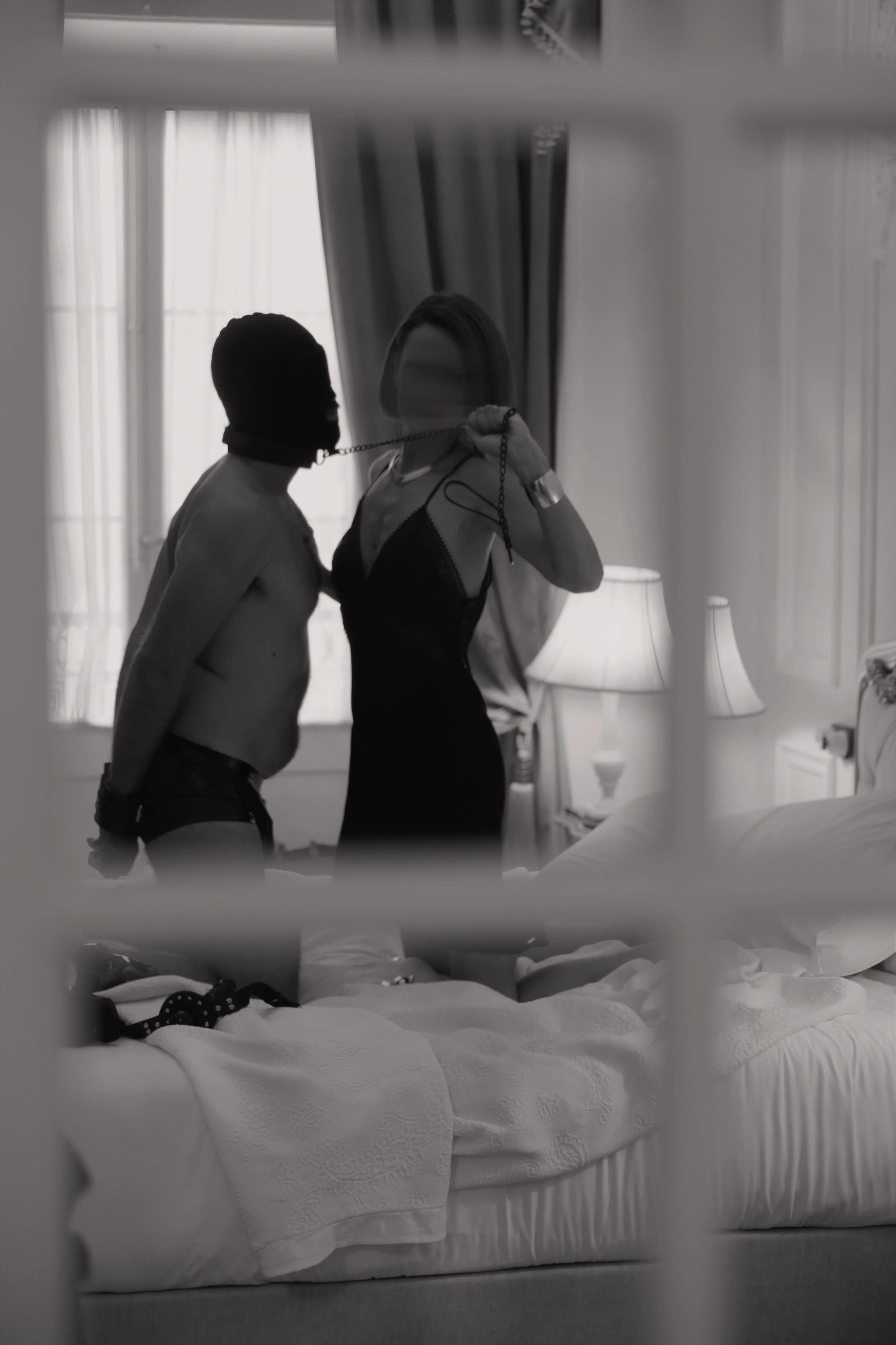 Mistress Devine, a leashed submissive man facing each other in front of a mirror in a bedroom.