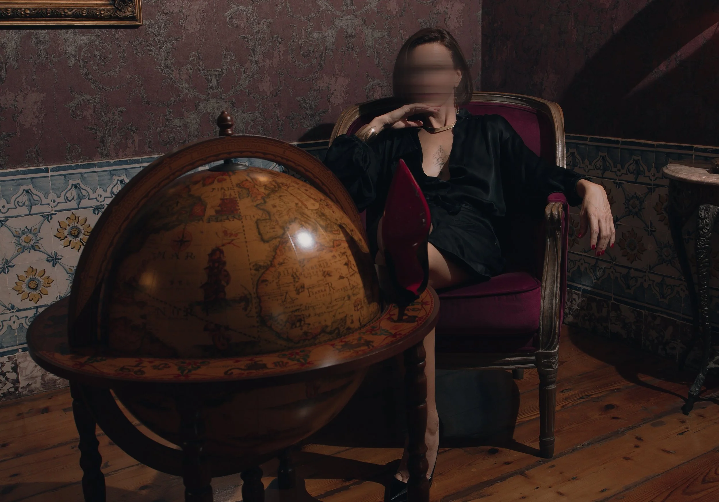 A dominant woman, Mistress Devine sitting in a vintage armchair, holding a red feather quill, with a large globe in front of her, a patterned wallpaper, a framed artwork on the wall, and a carved wooden side table in a dimly lit room. Red sole.