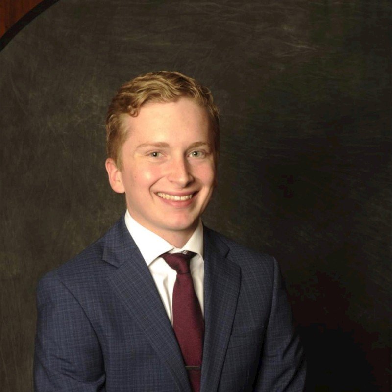 Portrait of a young man with light skin, curly blonde hair, dressed in a dark plaid suit, white shirt, and maroon tie, smiling against a dark backdrop.