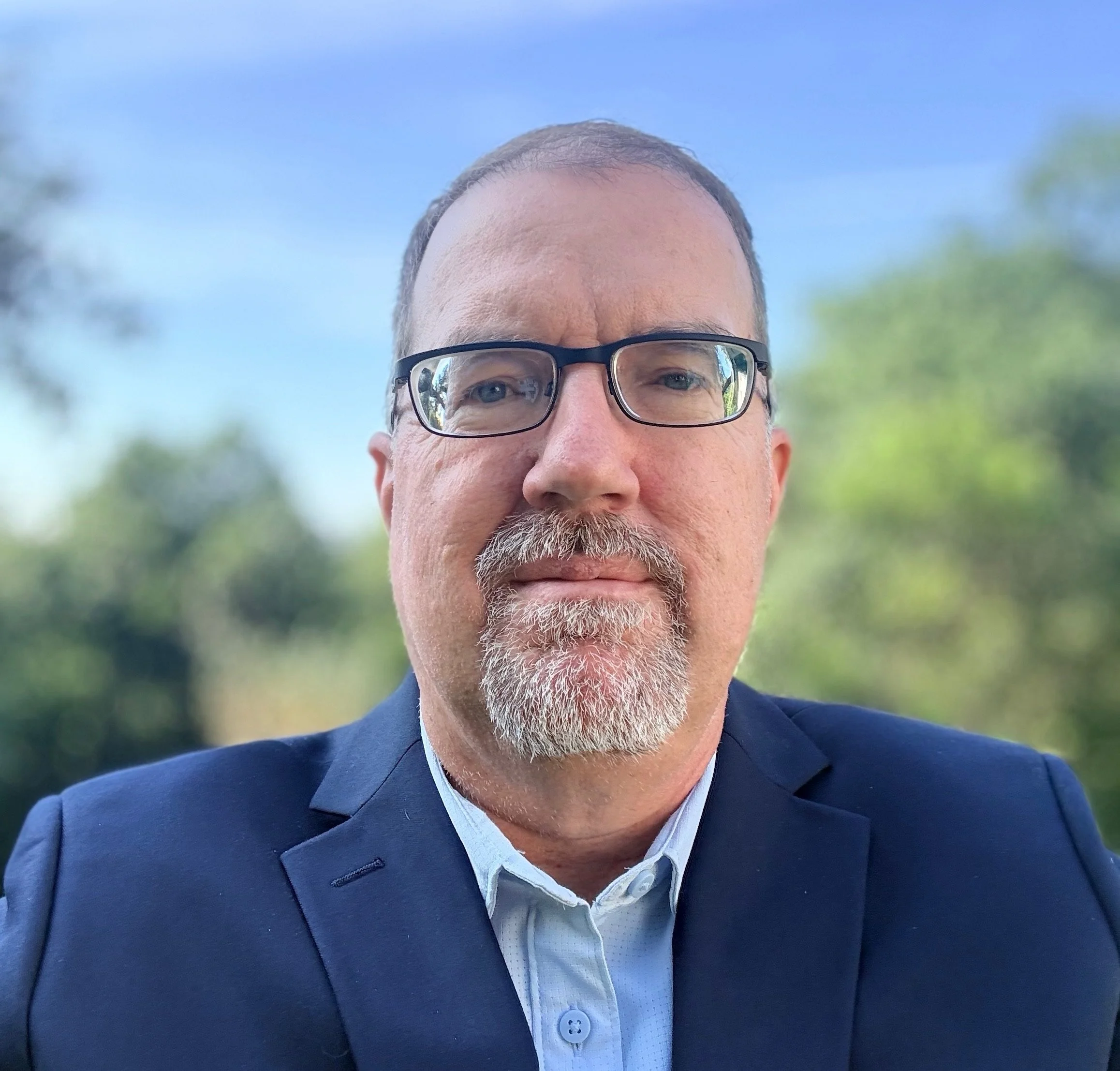 Close-up of a middle-aged man with light skin, gray hair, and glasses, wearing a suit and light blue shirt outdoors on a sunny day.