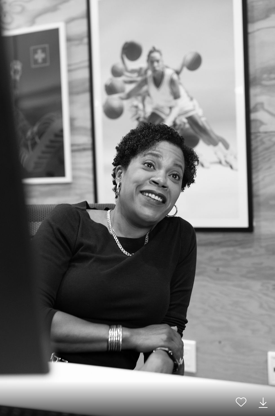 A woman with short curly hair smiling, seated at a table in a room with basketball-related posters on the wall behind her.