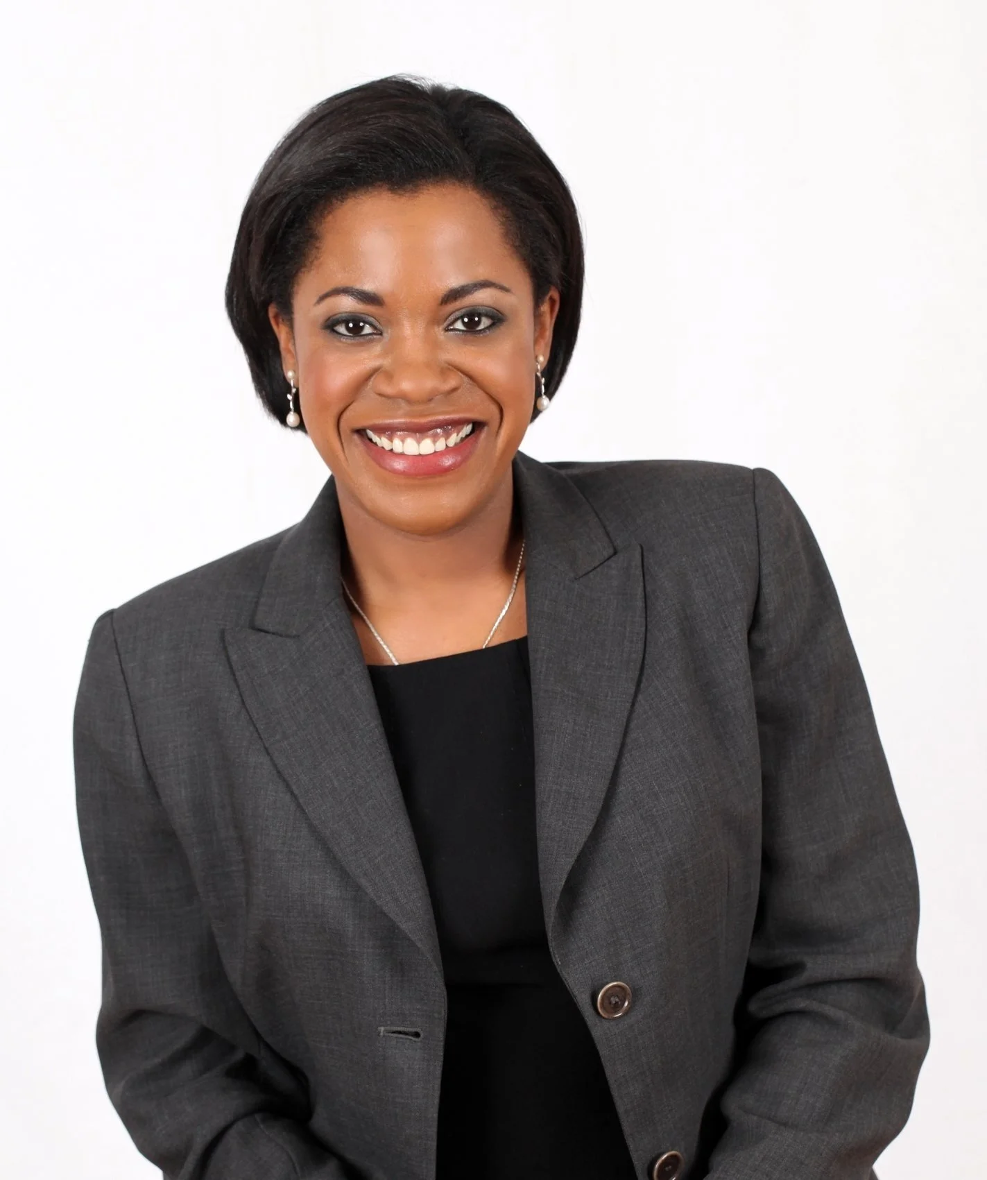 A smiling African American woman wearing a dark gray business suit with pearl earrings and a necklace, against a plain white background.