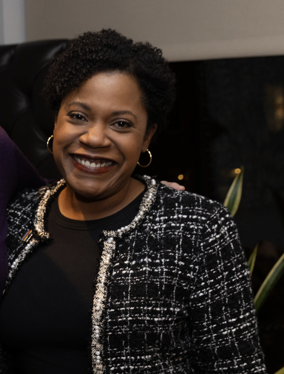 A smiling woman with short curly hair, wearing a black top and a black-and-white patterned jacket, sitting indoors at night.