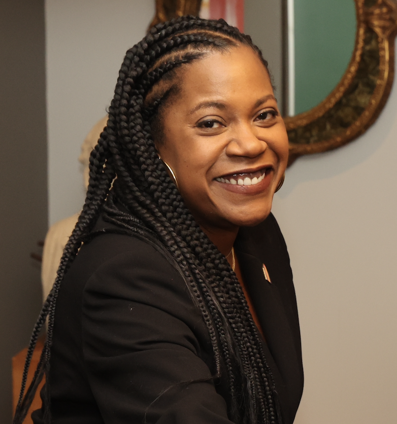 Smiling woman with braided hair, wearing a black blazer, in an indoor setting with a mirror in the background.
