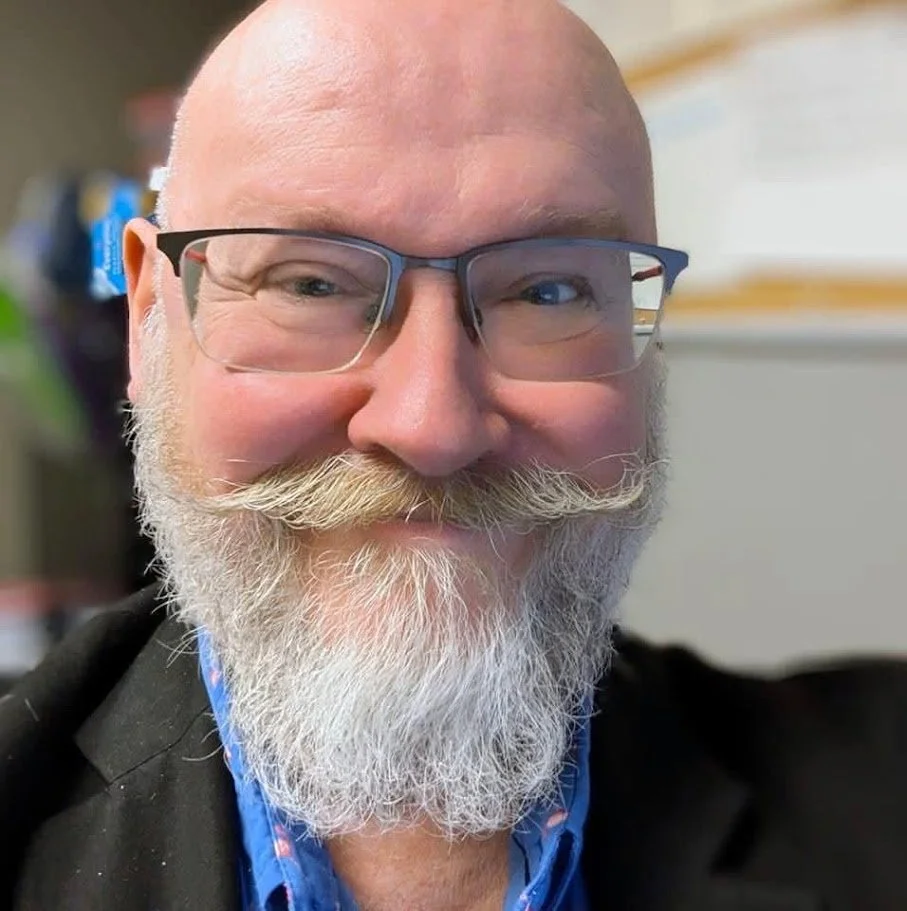 Close-up of a smiling bald man with a white beard and mustache, wearing glasses and a blue shirt.