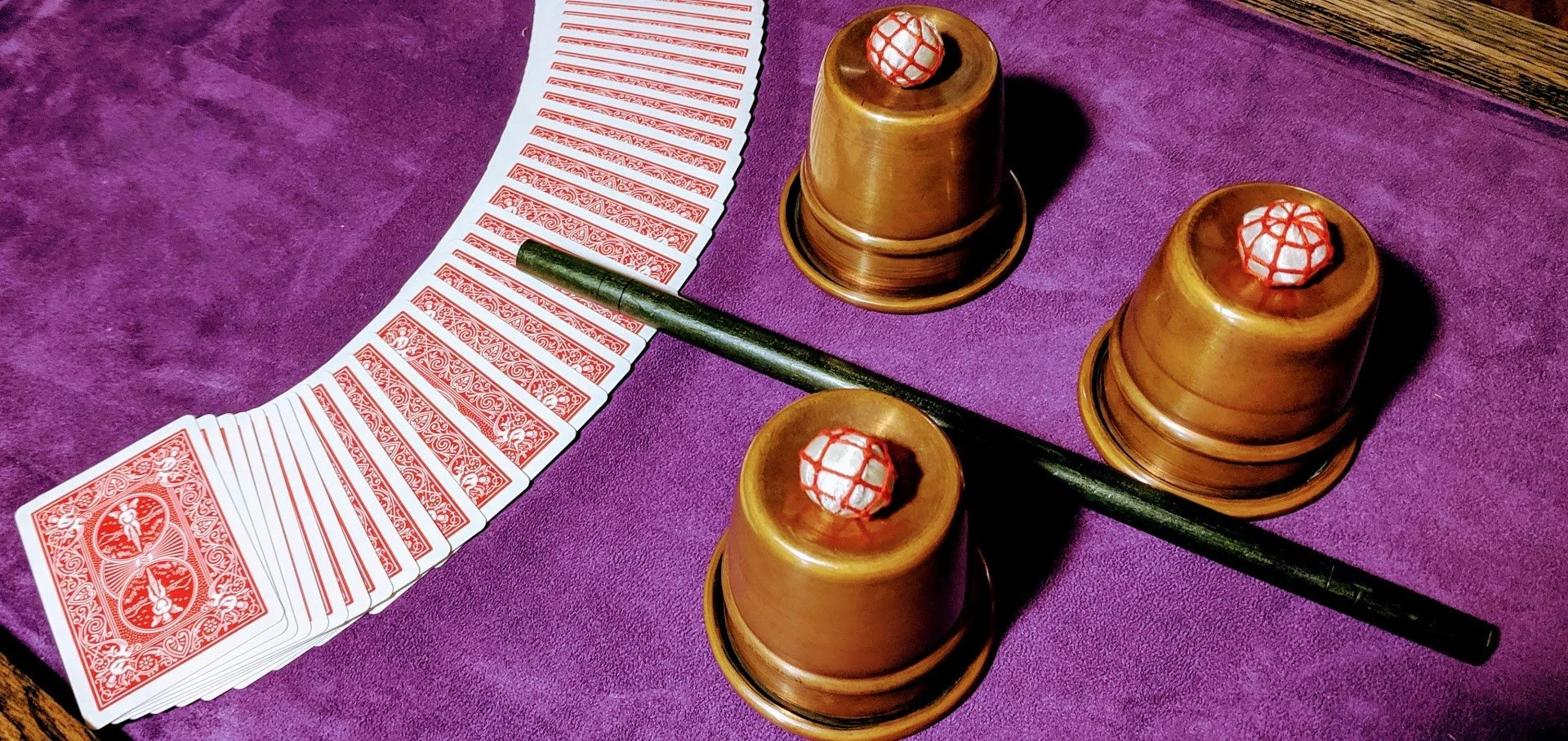 A game of Chinese Takeout, with a deck of red-backed playing cards fanned out on a purple felt surface. Three brass cups with red and white balls inside are arranged in a triangular formation, with a black stick passing across and resting on two cups.