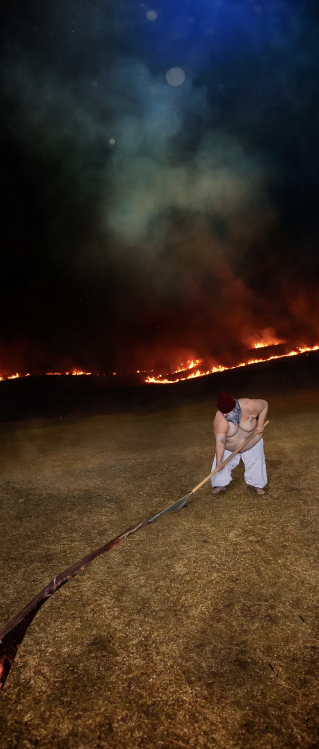 Person using a long-handled shovel near a nighttime grass fire with smoke and flames