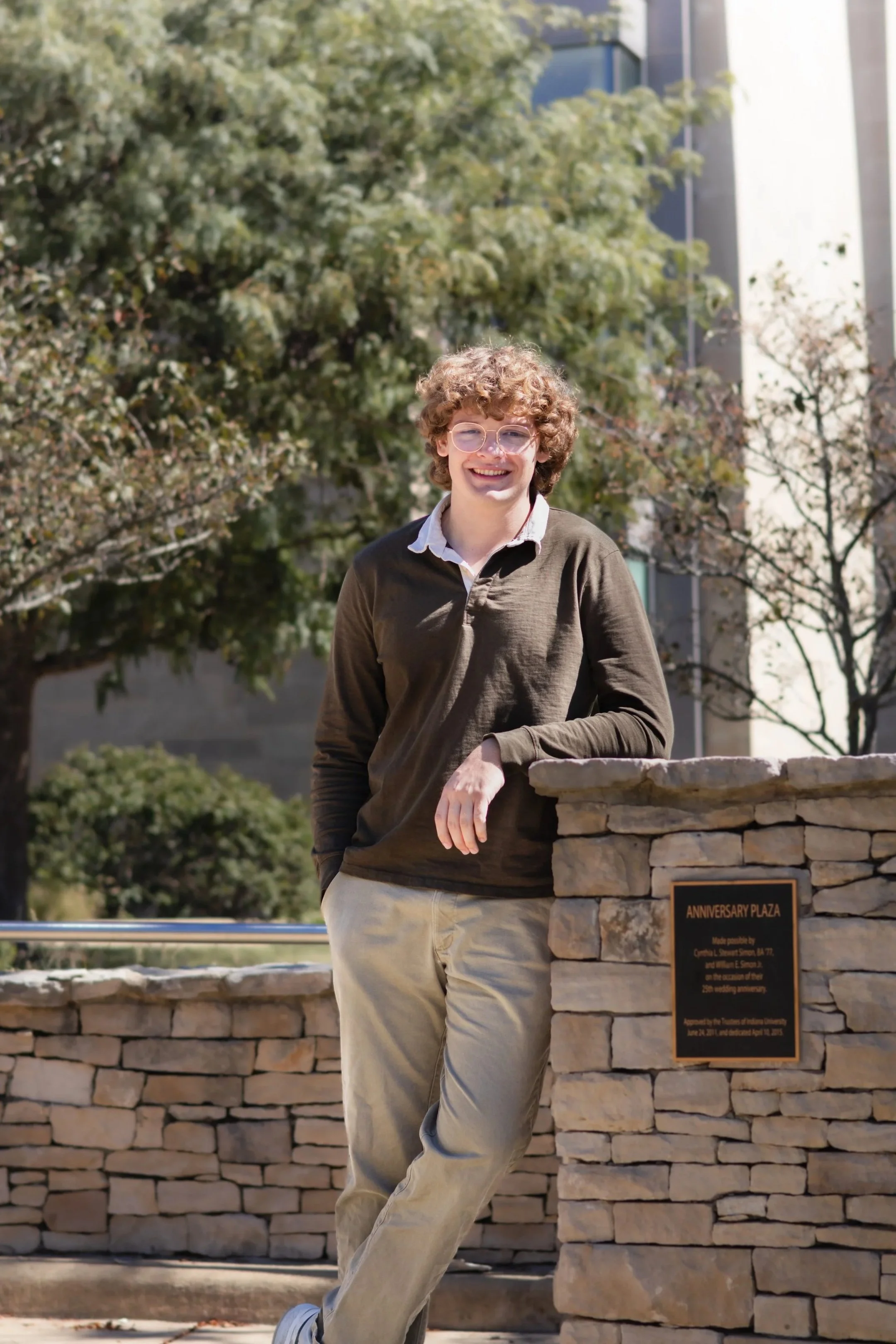 Smiling young man with curly hair and glasses standing beside a stone wall and Anniversary Plaza plaque outdoors