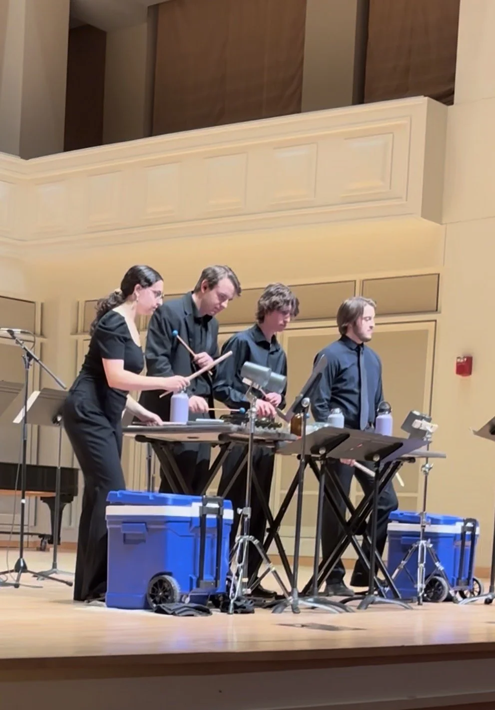 Four musicians performing with percussion instruments on a concert hall stage.