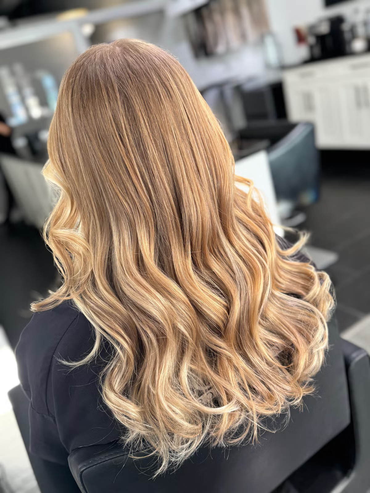 Back view of a woman with long, wavy, blonde hair in a salon setting. balayage