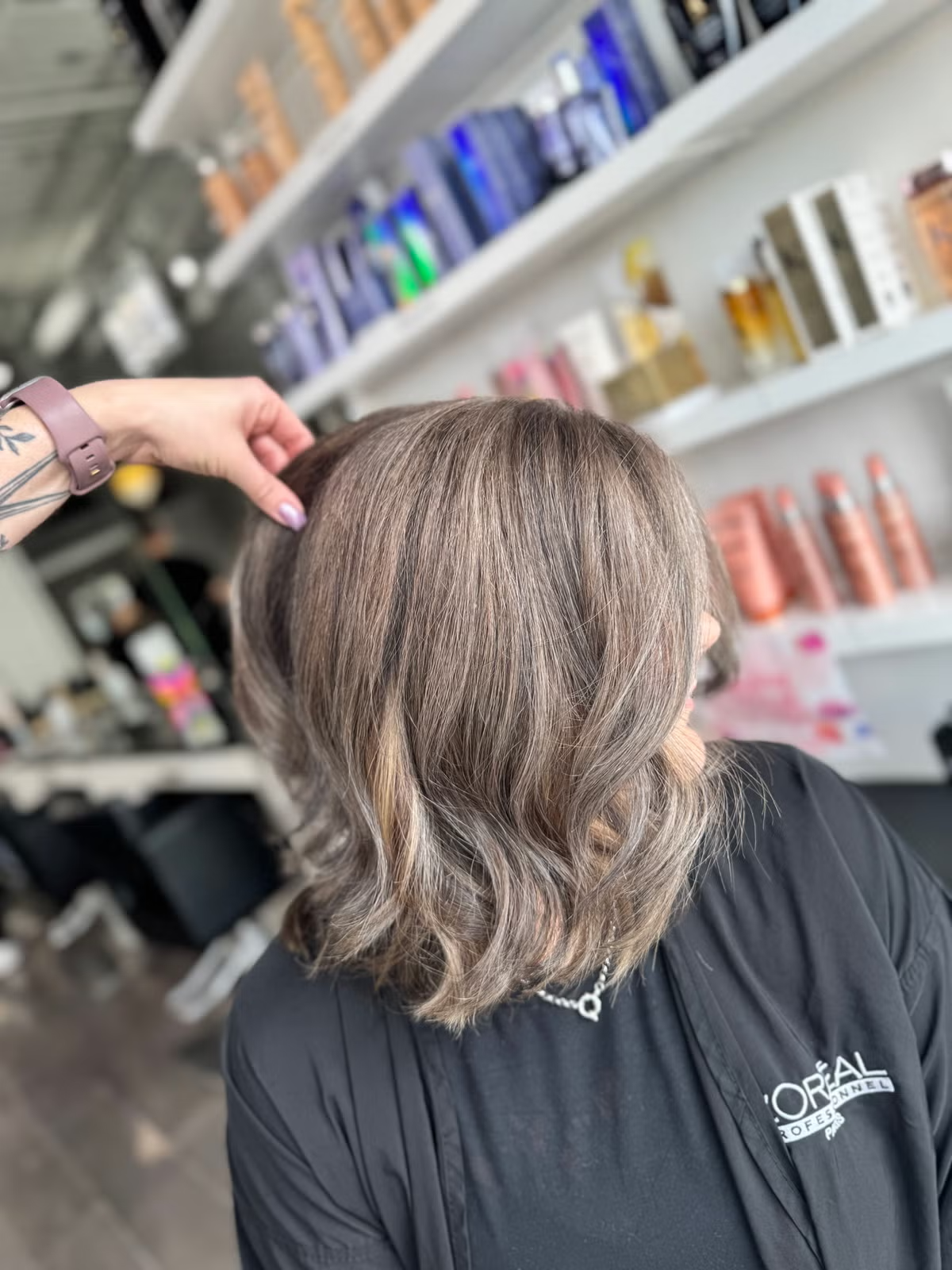 A woman with shoulder-length wavy hair with balayage highlights being styled at a hair salon, wearing a L'Oréal Professional apron.