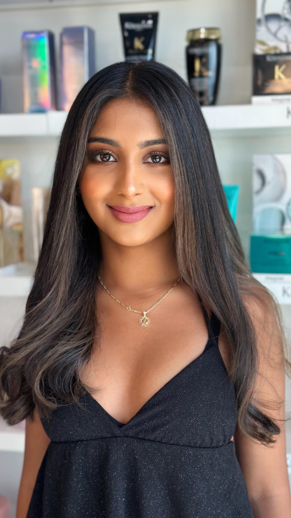 A young woman with long dark hair, wearing a black sleeveless dress and a gold necklace, standing in front of shelves displaying various beauty products.