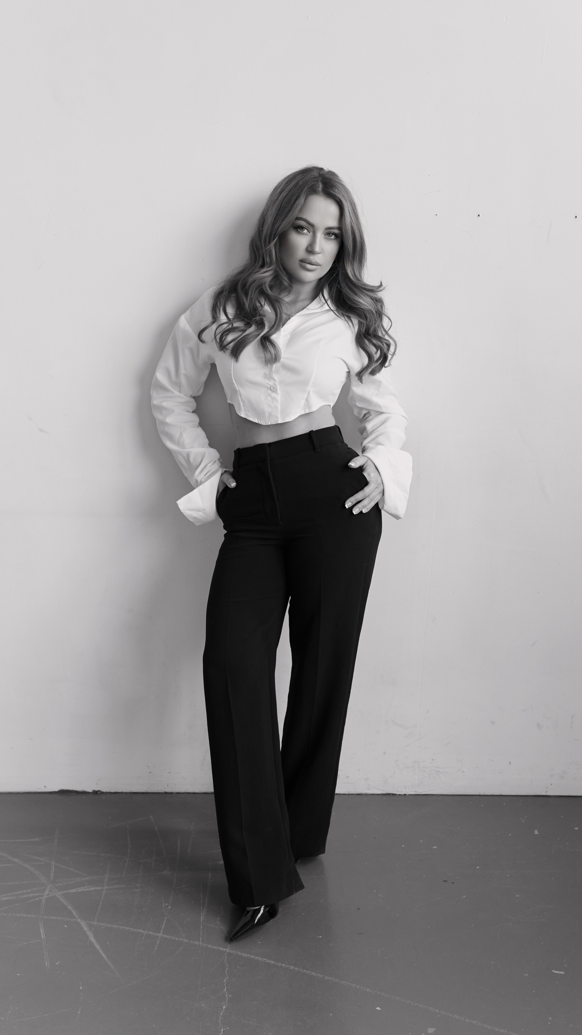 Black and white photo of a woman standing against a plain wall. She has long wavy hair and is wearing a cropped button-up shirt and high-waisted pants, with one hand on her hip and the other in her pocket.