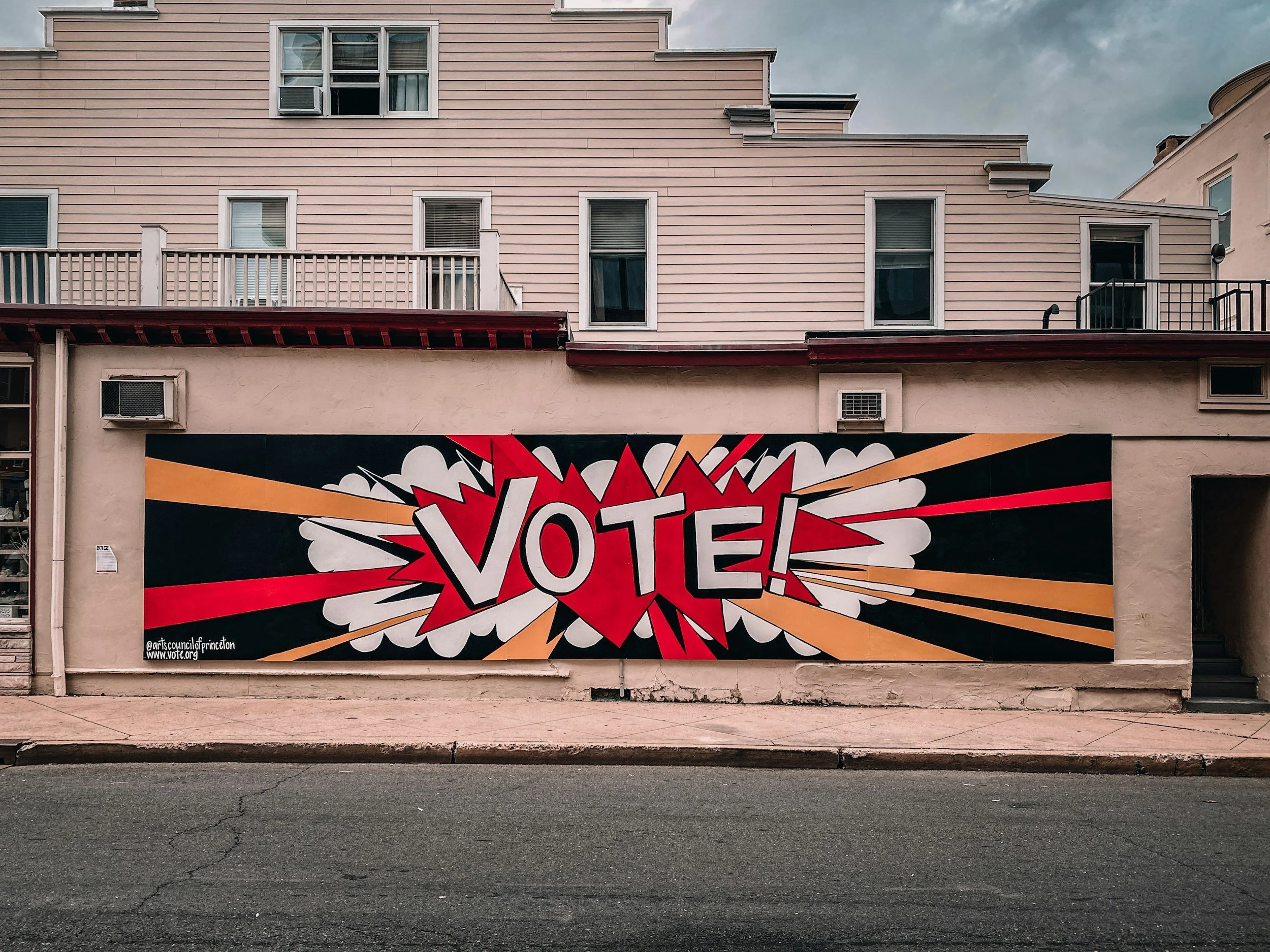 Street-level mural with a bold, colorful comic book style design featuring the word "VOTE!" in large, red, white, and black letters with explosion effects and accompanying graphics, painted on the side of a building under a cloudy sky.