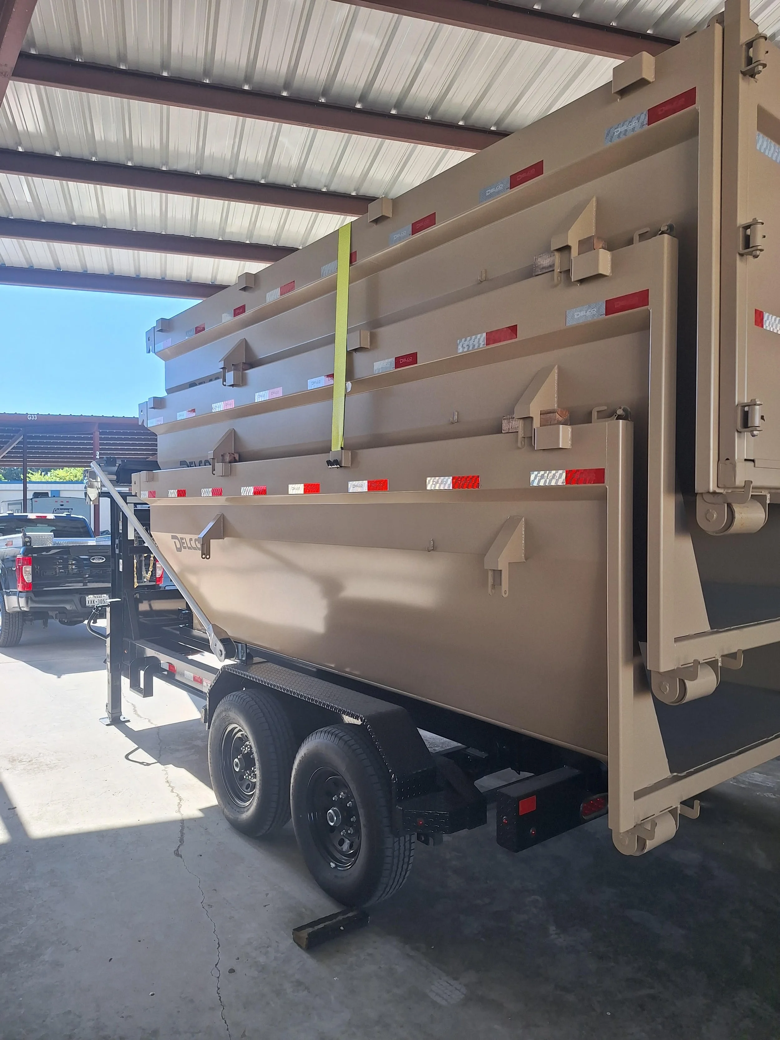 A large beige utility truck trailer parked under a metal roof, with multiple stacked compartments and a yellow strap hanging from the top. The background shows pickups and a clear blue sky.