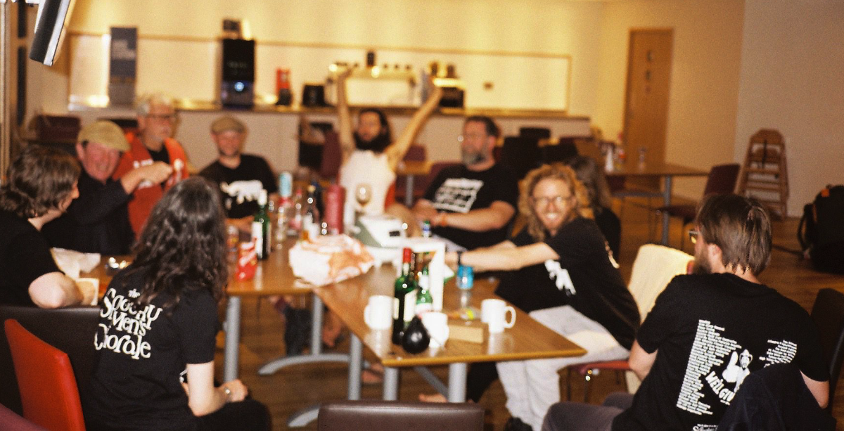 Group of people sitting around a table in a casual setting, some smiling and raising their hands, with food, drinks, and mugs on the table.