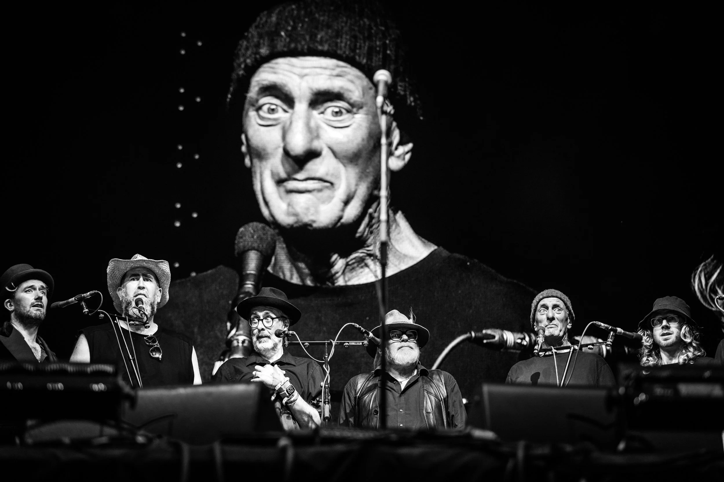 Black and white image of a group of men on stage, with a large screen behind them displaying a close-up of a man's face. The men are wearing hats and glasses, and appear to be singing or performing.