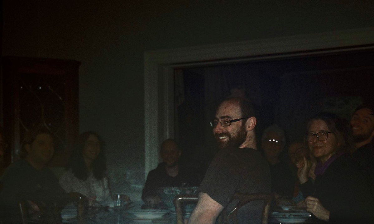Group of people gathered around a table in a dimly lit room, smiling and looking towards the camera.