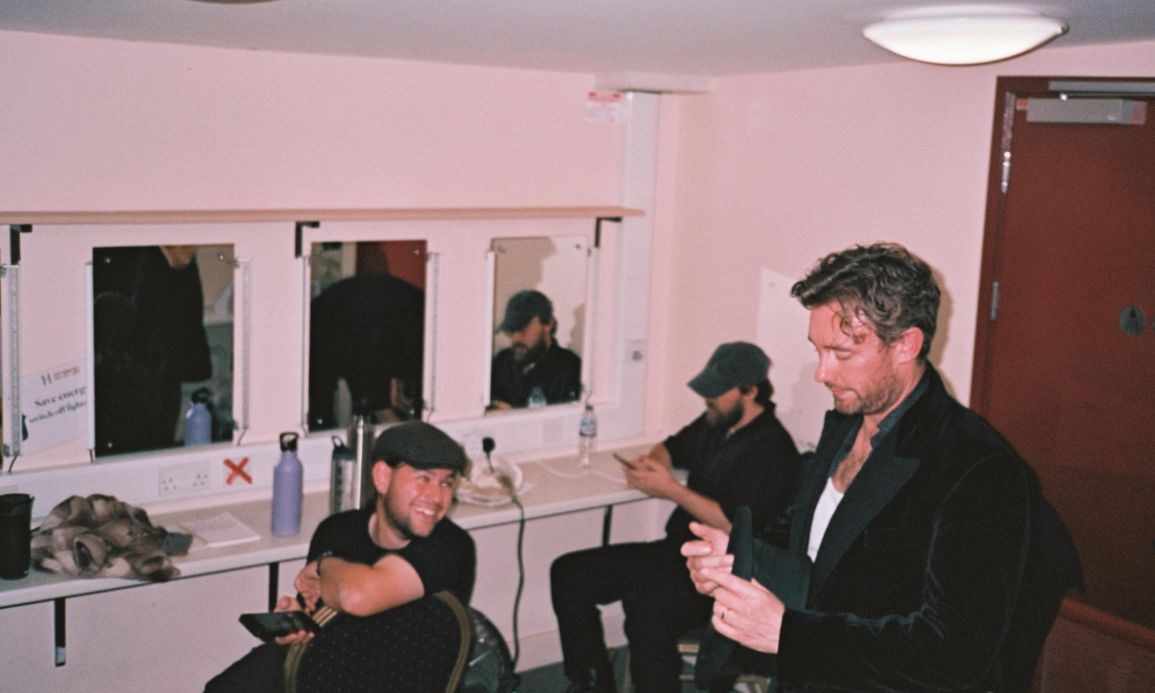 Four men in a dressing room, three sitting at a counter with mirrors, one standing. One man is smiling and holding a phone, another is looking at his phone, and the other two are engaged with their phones. The room has pink walls, mirrors, and variou