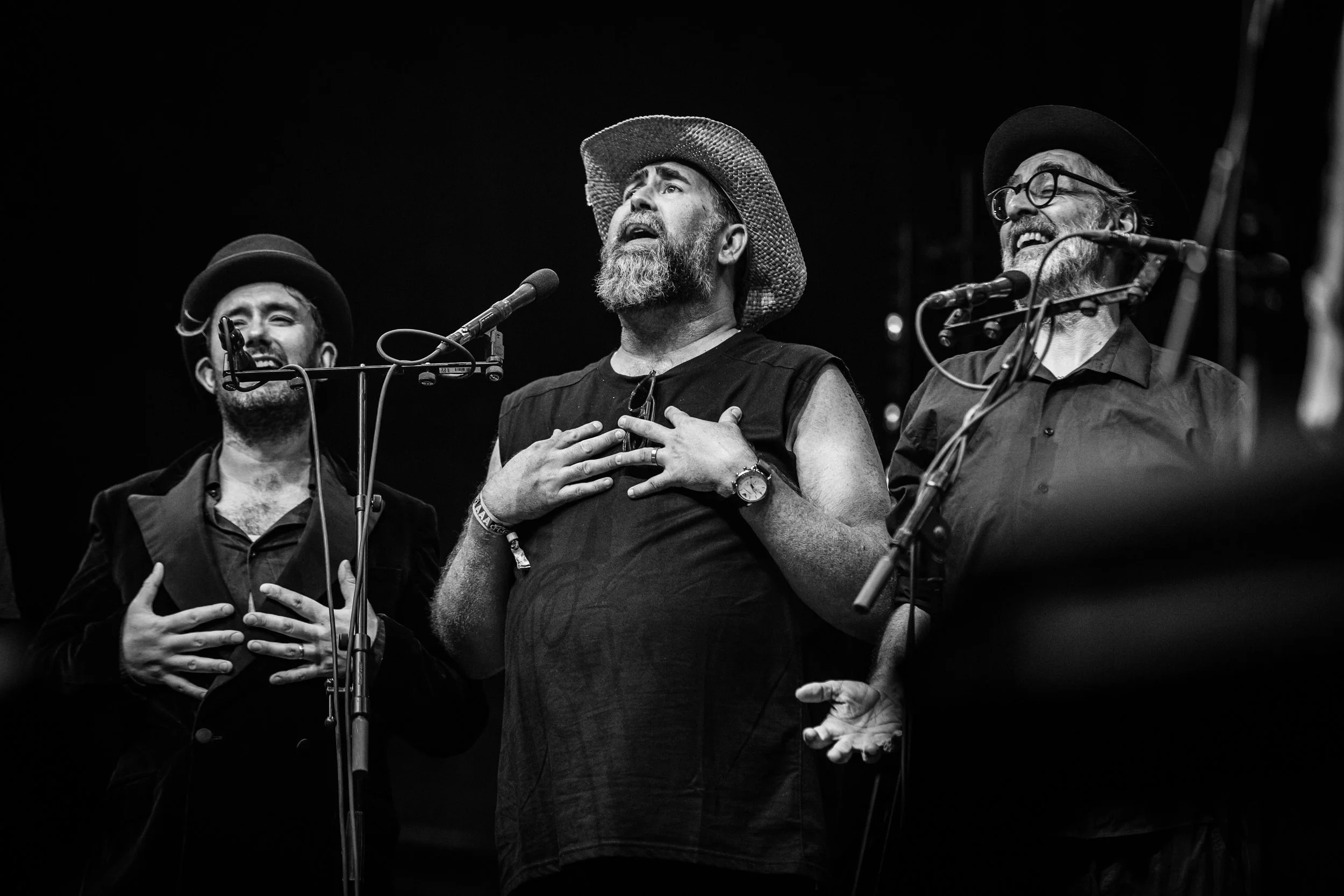 Three men singing on stage, wearing hats, with one man holding his chest in front of a microphone, in black and white.