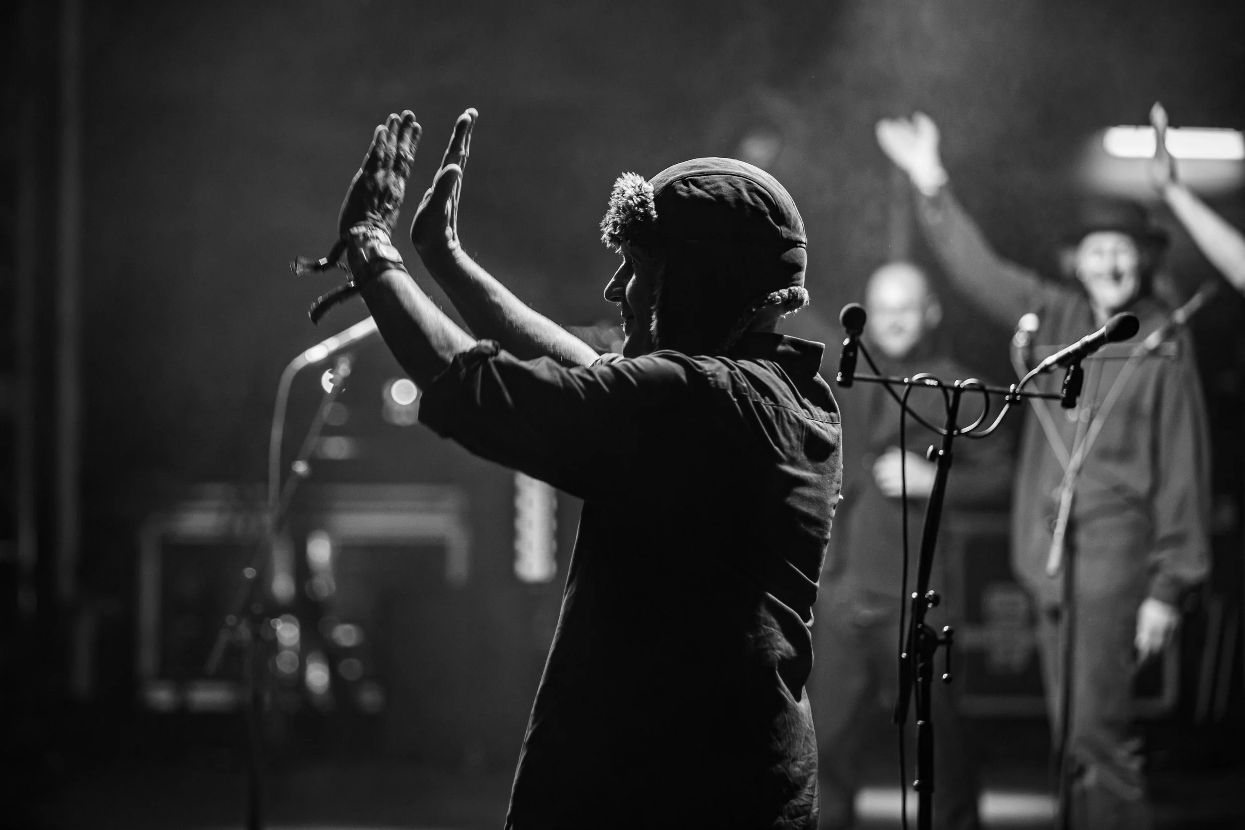 Black and white photo of a pianist playing piano on stage, wearing a hat with ear flaps and a jacket, with microphones nearby and a person in background raising a hand.