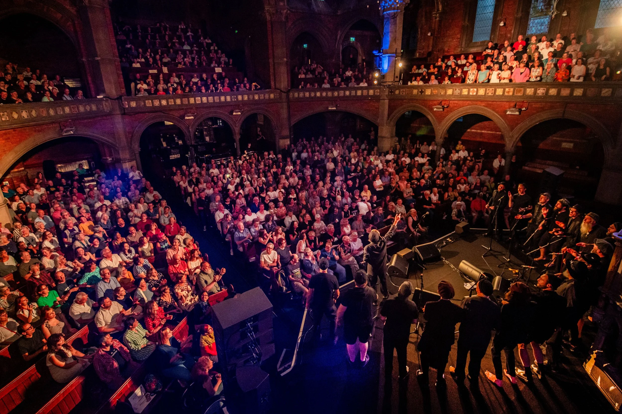 A large audience seated in a tiered concert hall listening to a performance on stage. The hall features arched architecture and large windows, with colorful lighting illuminating the space.