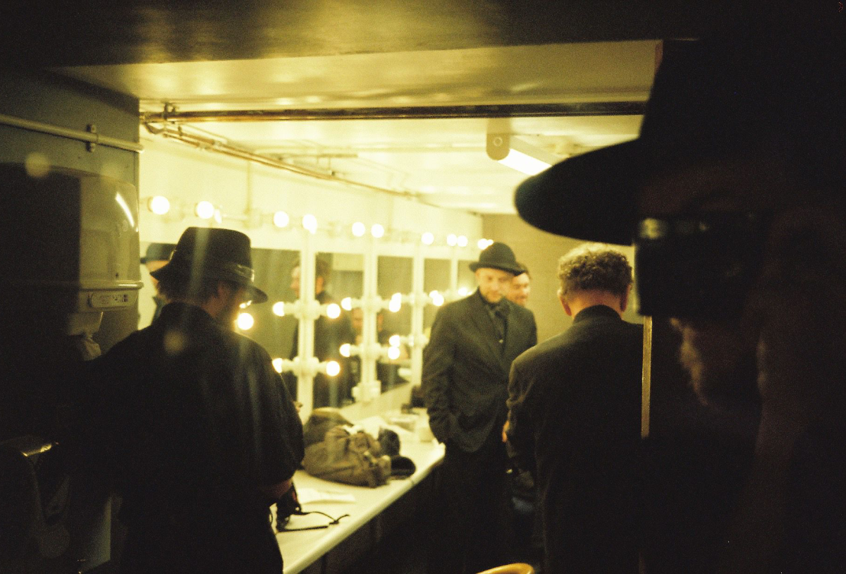 People in dark clothing and hats get ready behind the scenes in a dressing room with bright mirror lights.