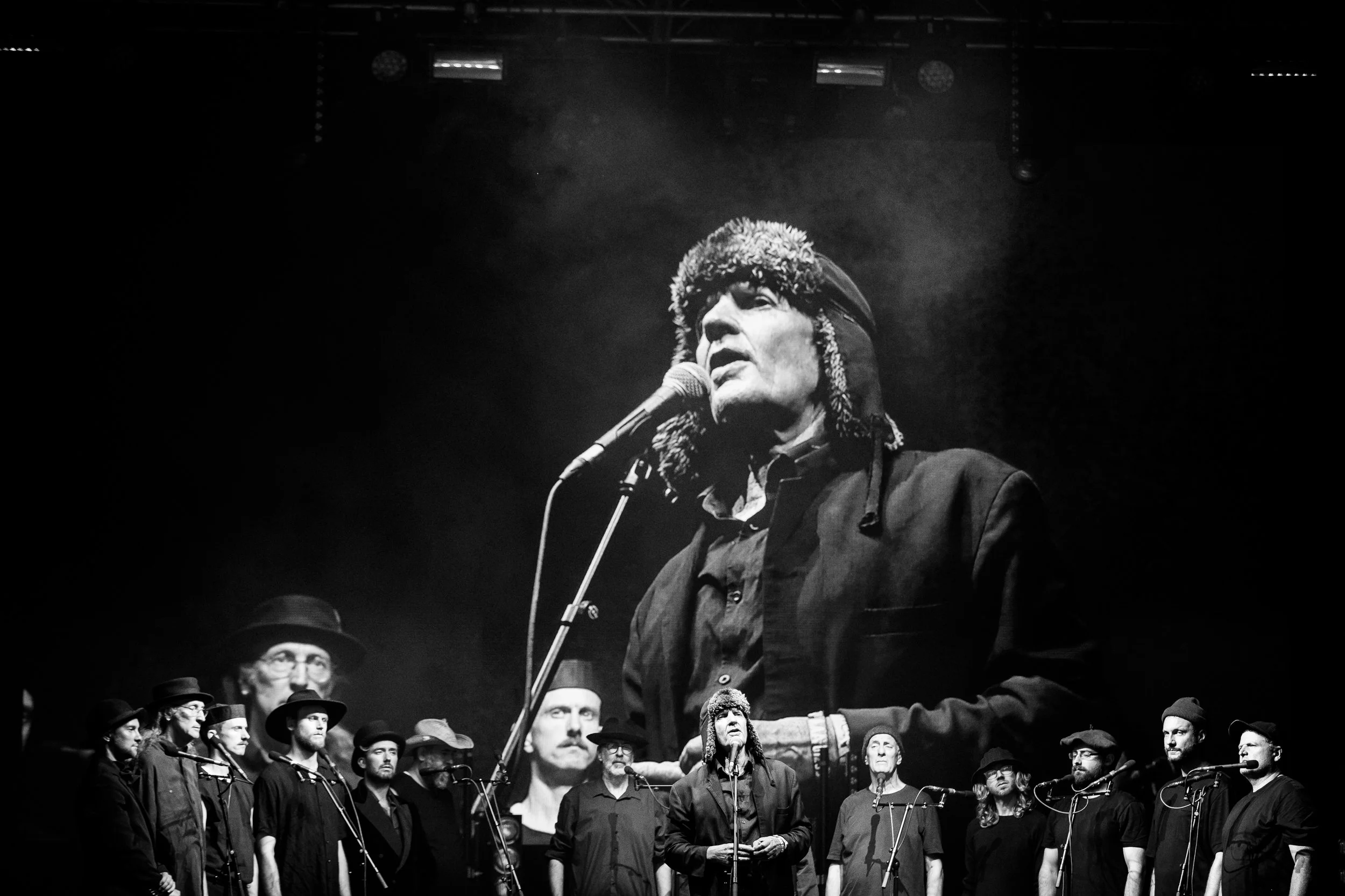 A black and white photograph of a man wearing a furry hat and dark jacket speaking or singing into a microphone on stage, with a group of people standing behind him, some wearing hats, in a dimly lit setting.
