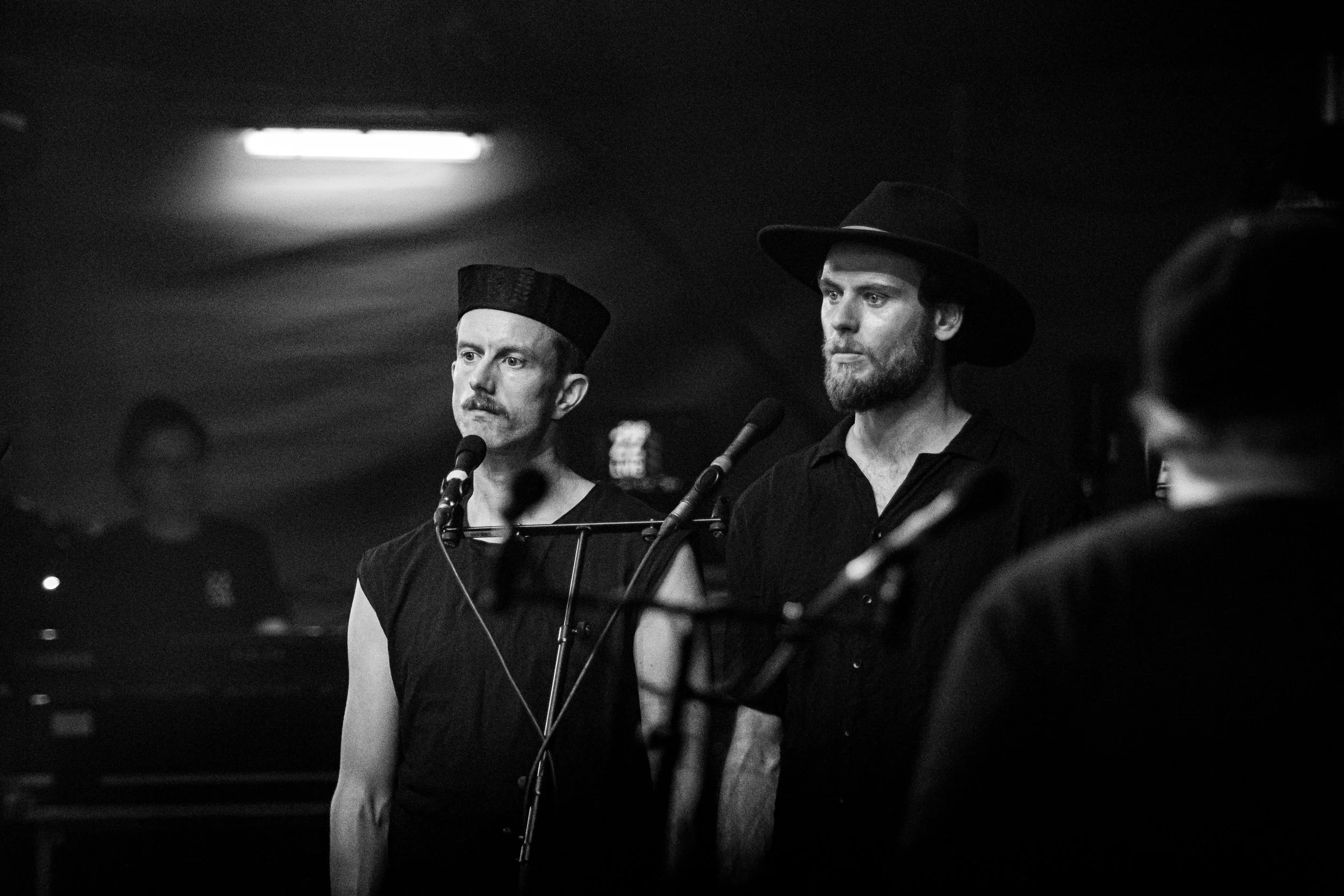 Black and white photo of two men singing into microphones; one wears a hat, the other a traditional hat, with a dark background.