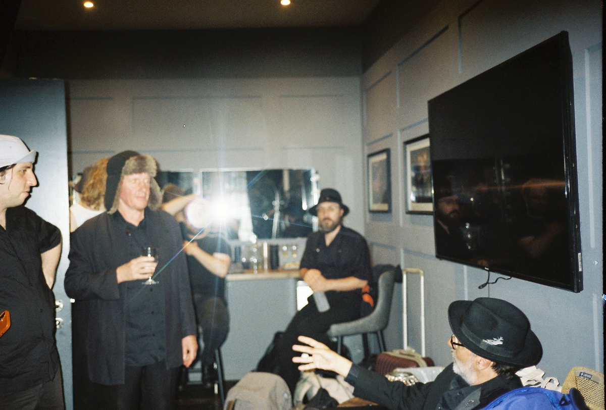 Group of people at a social gathering in a room with framed pictures on the wall, a large TV, and a mirror, some holding drinks, one person reaching out, and another sitting on a stool wearing a black hat.