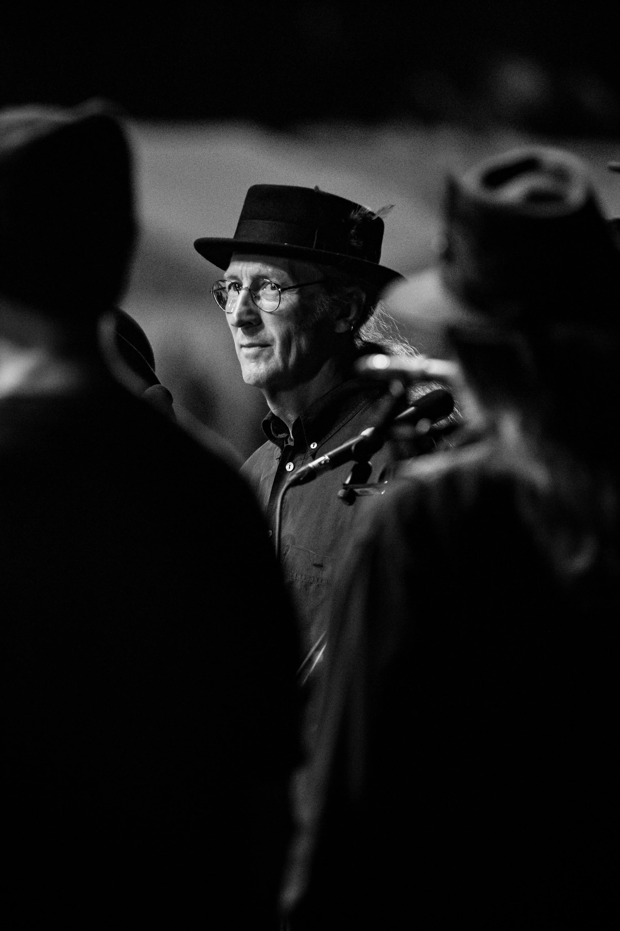 Black and white photograph of a man wearing glasses and a top hat, surrounded by blurred figures, possibly in a social or performance setting.