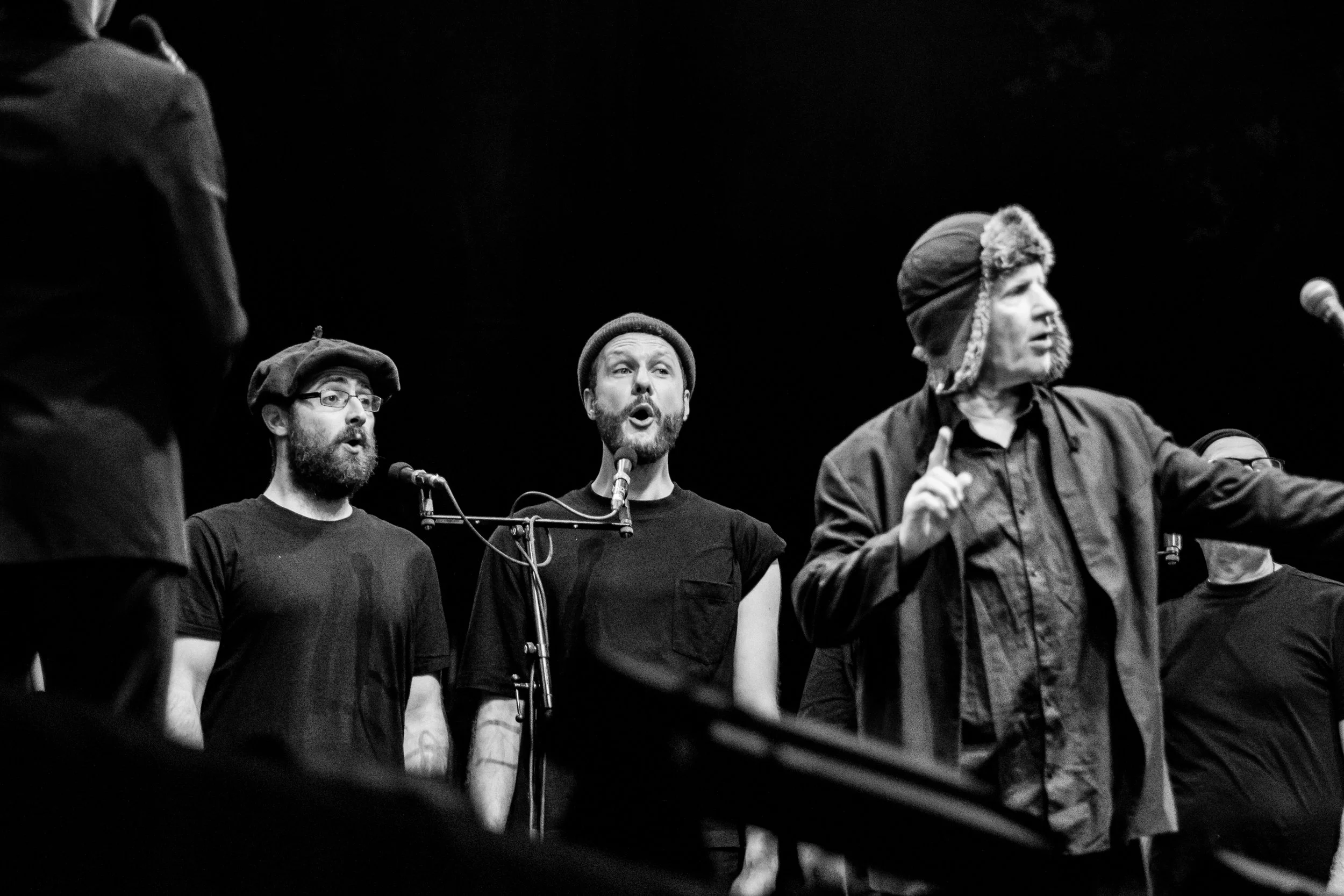 Black and white photo of four men singing into microphones on a stage, with dark background.