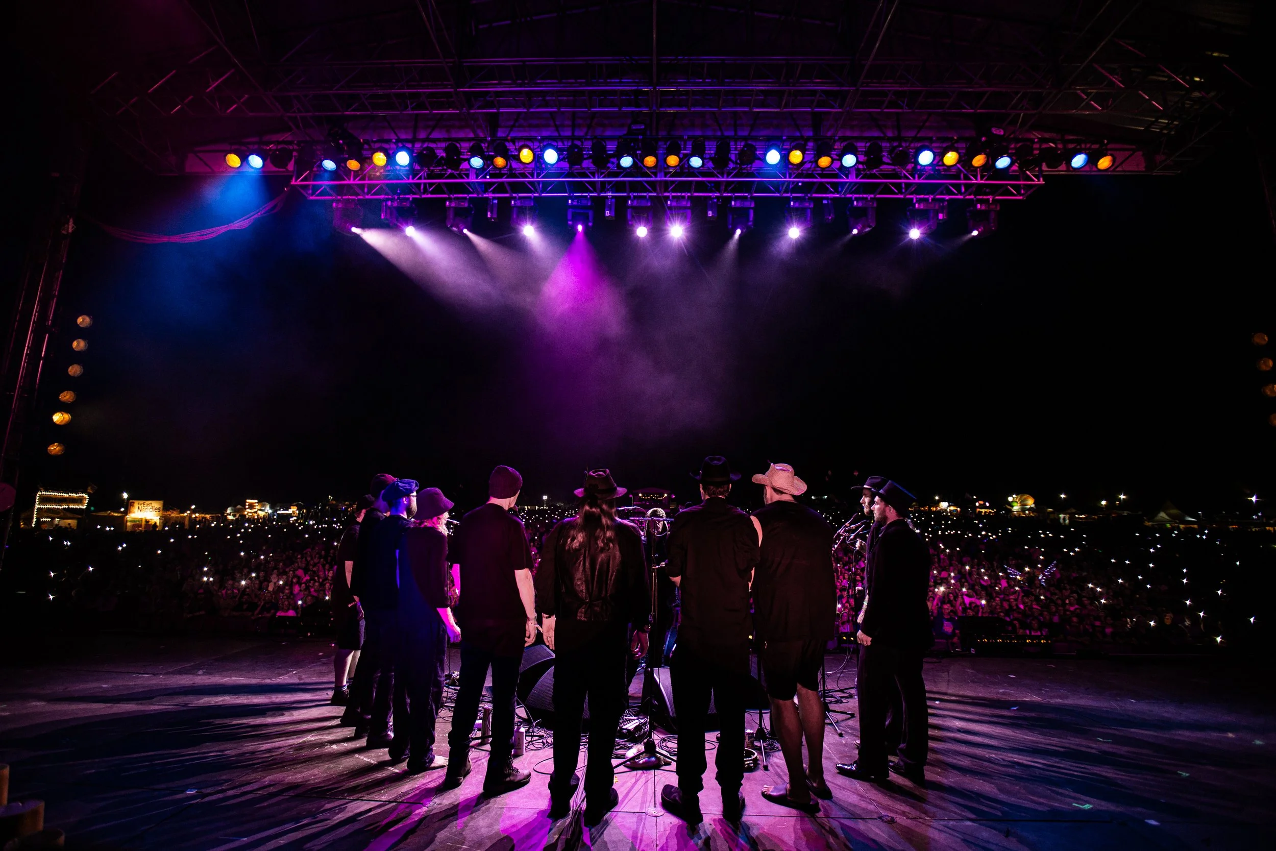 A group of people standing on a stage at night, watching a concert with colorful stage lighting and a large crowd in the background.