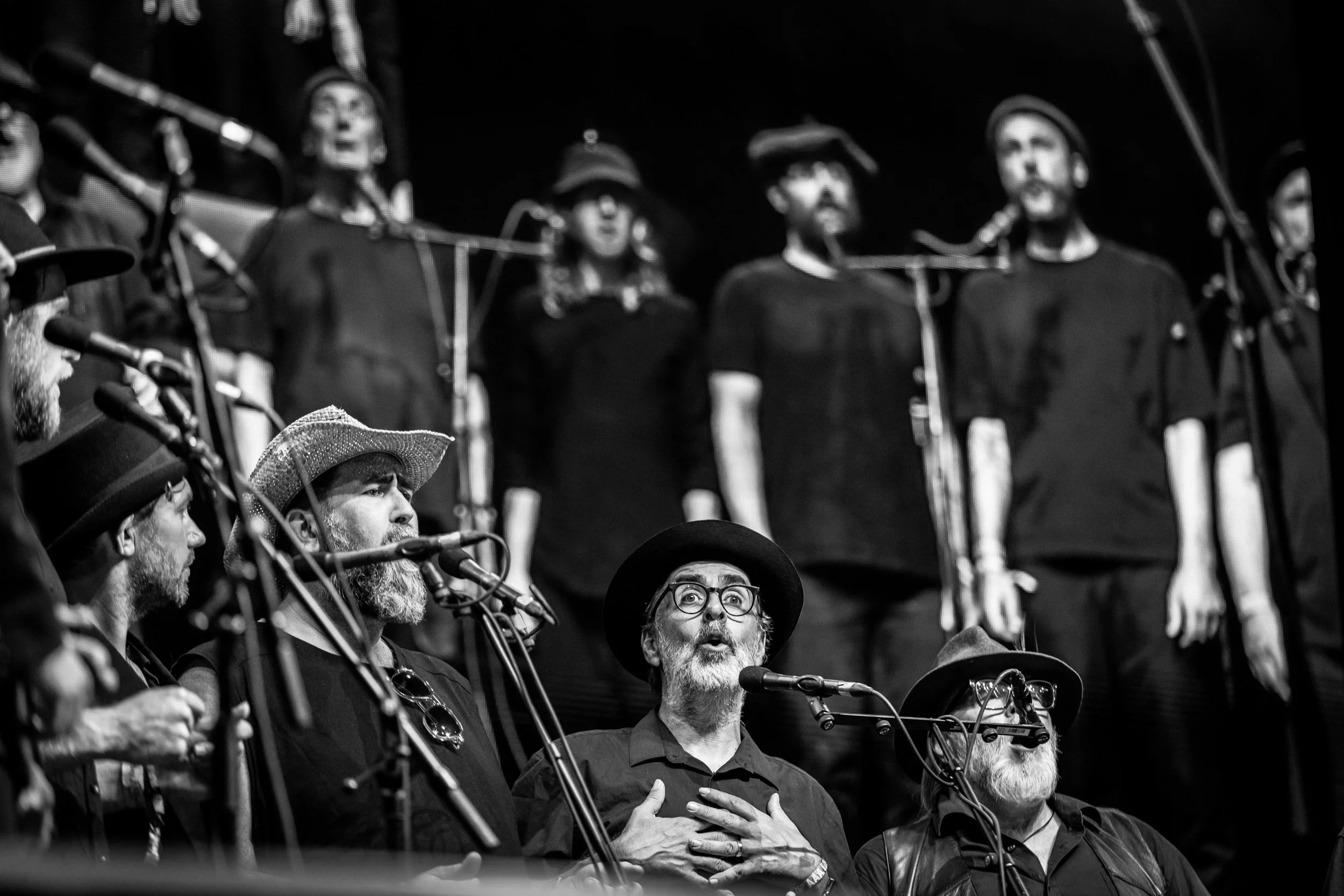 A black and white photo of a choir performing on stage, with several singers standing and some playing instruments, in front of microphones.