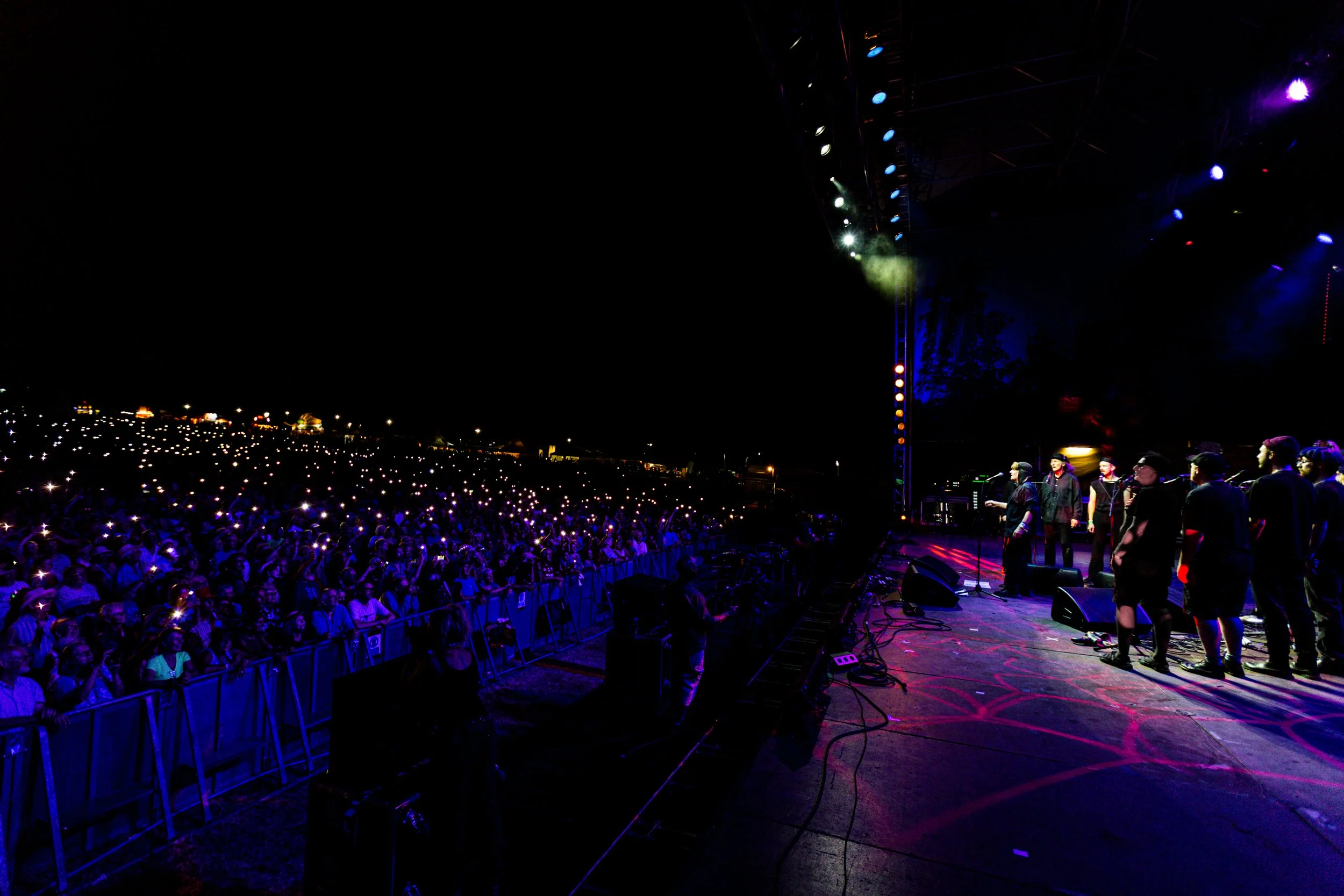 Nighttime outdoor concert scene with a large crowd, many illuminated by phone lights, and performers on stage under colorful stage lights.
