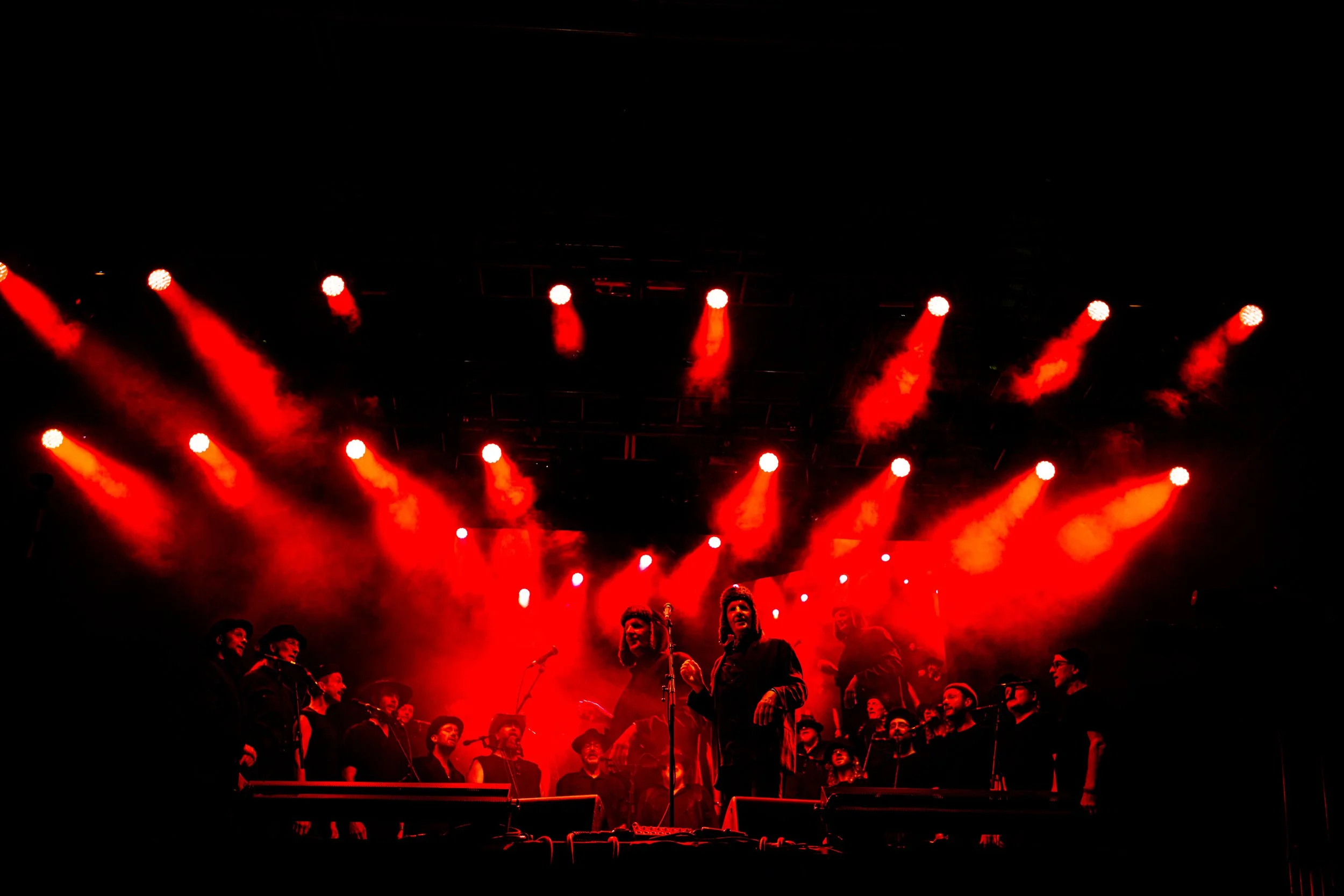 A concert stage illuminated with red lights, with a group of singers or performers standing in front of microphones, singing or speaking to an audience.