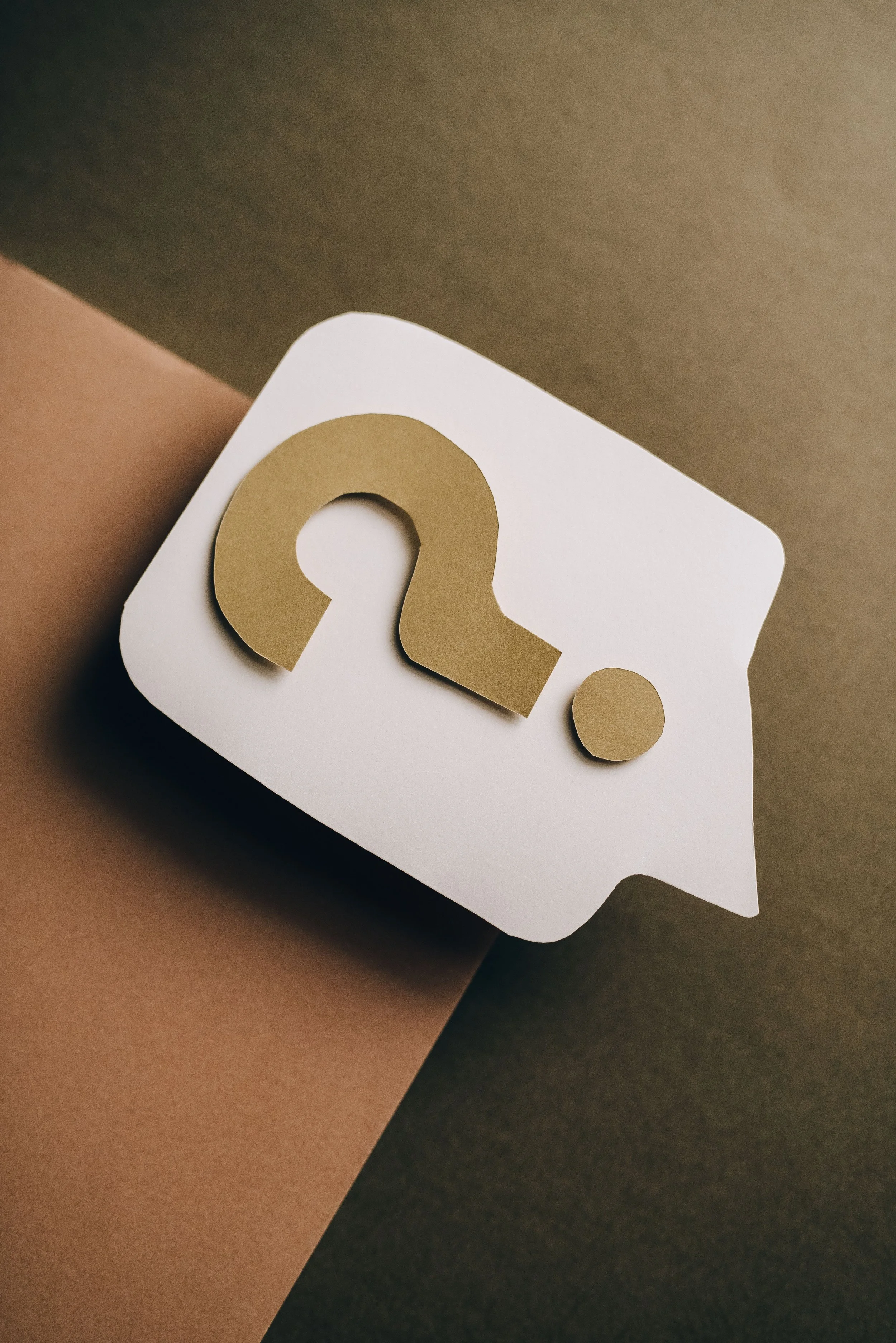 Une boîte blanche avec un symbole de point d'interrogation doré sur le dessus, placée sur des papiers de couleur beige, rose et gris foncé.