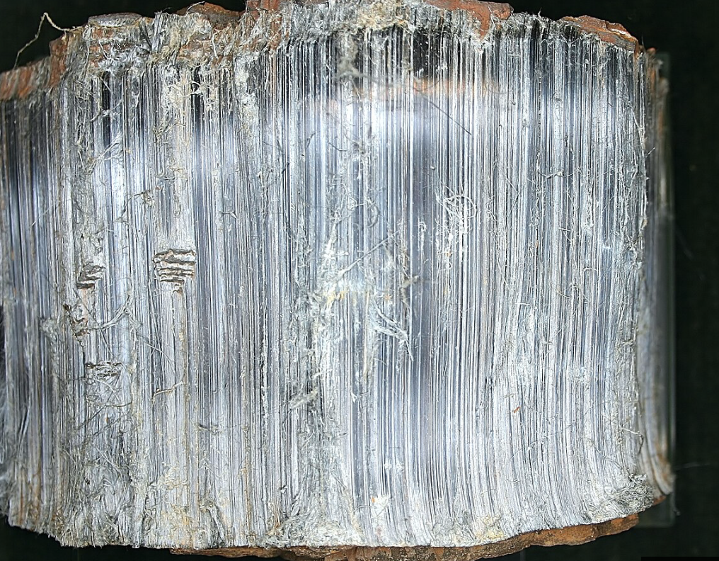 Close-up view of a metallic mineral specimen showing a fibrous, layered structure with a shiny, reflective surface and some rough, earthy edges.