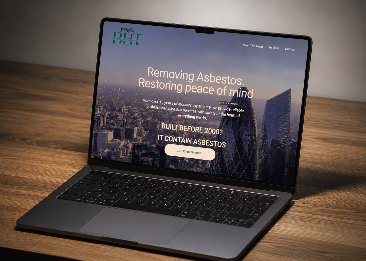A laptop on a wooden desk displaying a website for a company specializing in asbestos removal. The website features a city skyline background with text about removing asbestos and restoring peace of mind.