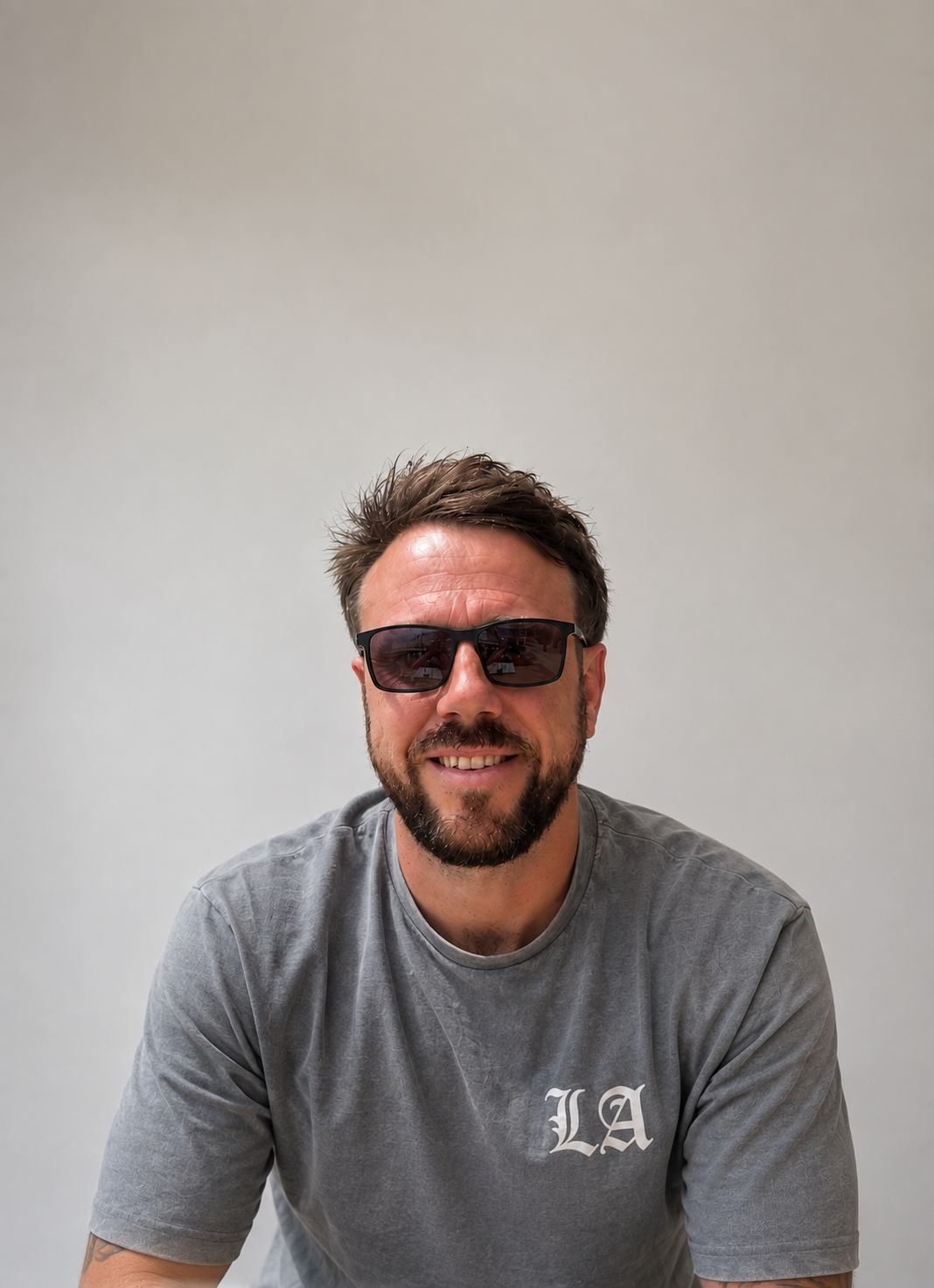 A smiling man with a beard, wearing sunglasses and a gray T-shirt with 'LA' on it, against a plain white background.