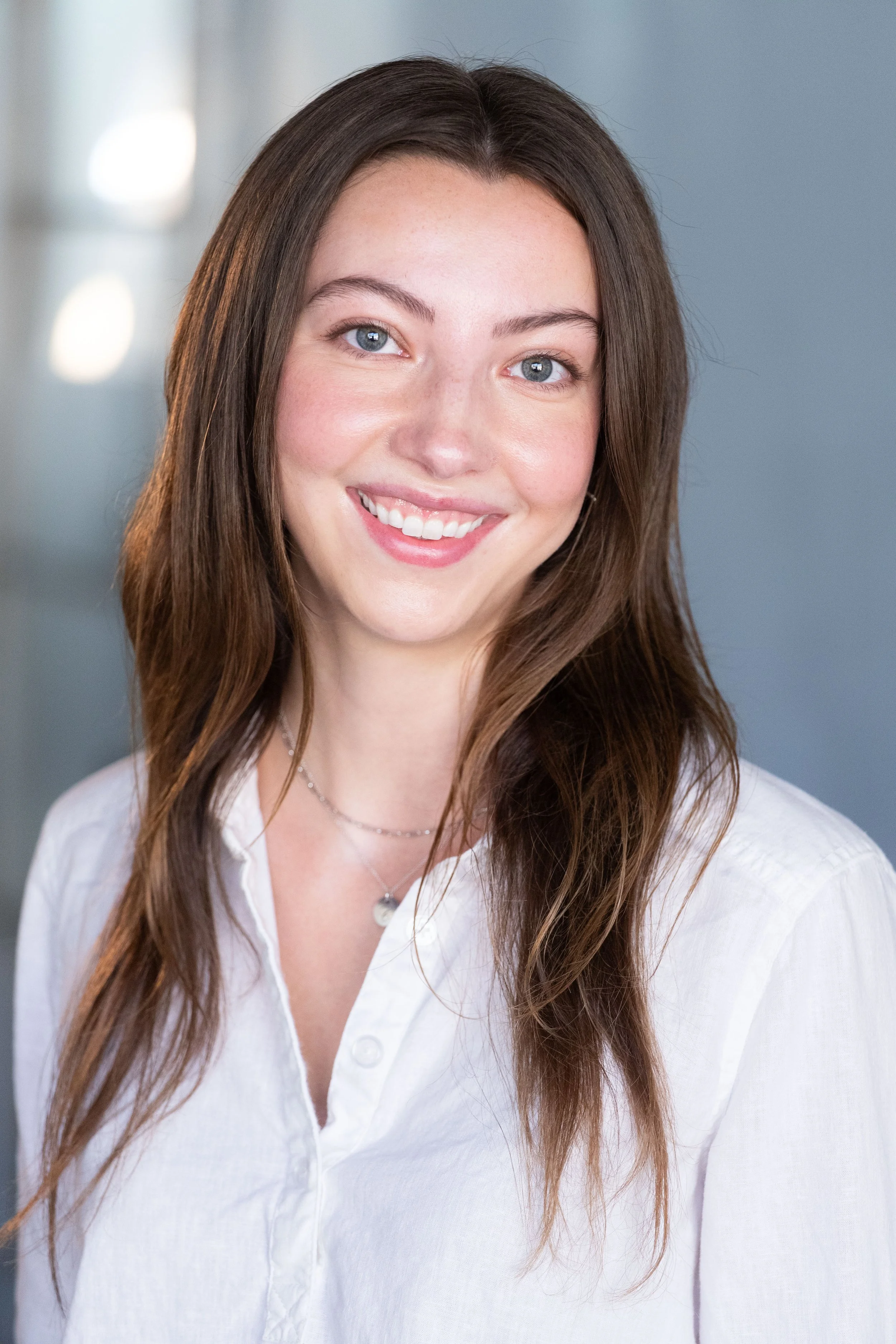 A young woman with long brown hair and blue eyes smiling, wearing a white shirt and a silver necklace, standing indoors with a blurred background.