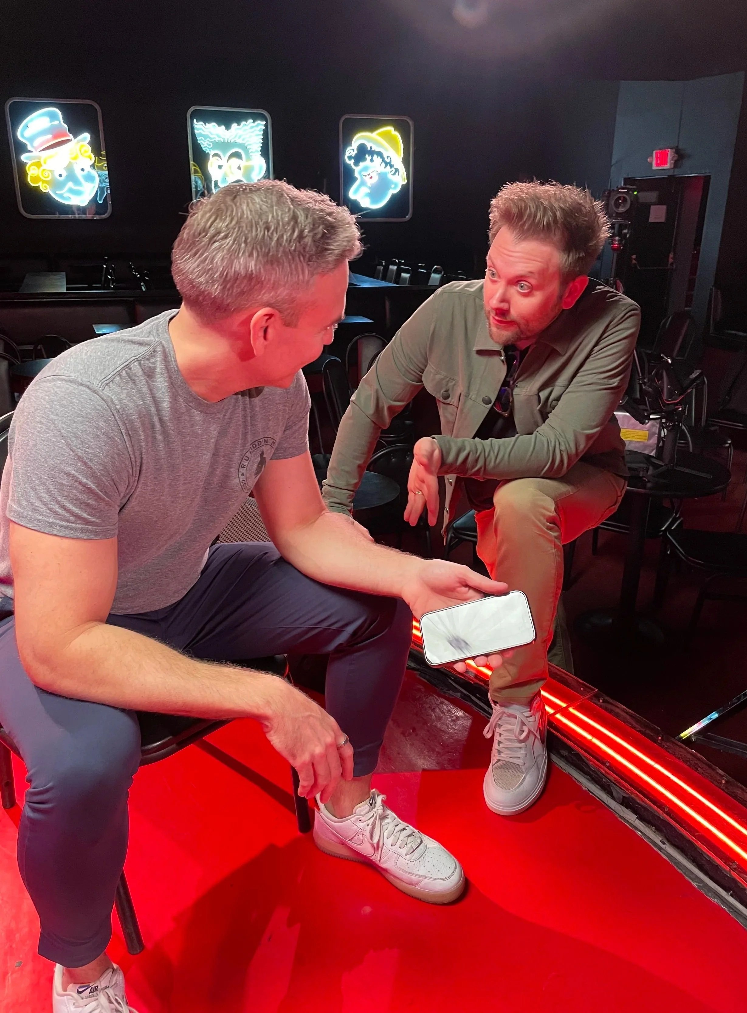 Two men sitting on black chairs, one showing his leg to the other while the latter points at his shoe, inside a dimly lit room with colorful neon signs of clown faces on the wall behind them.
