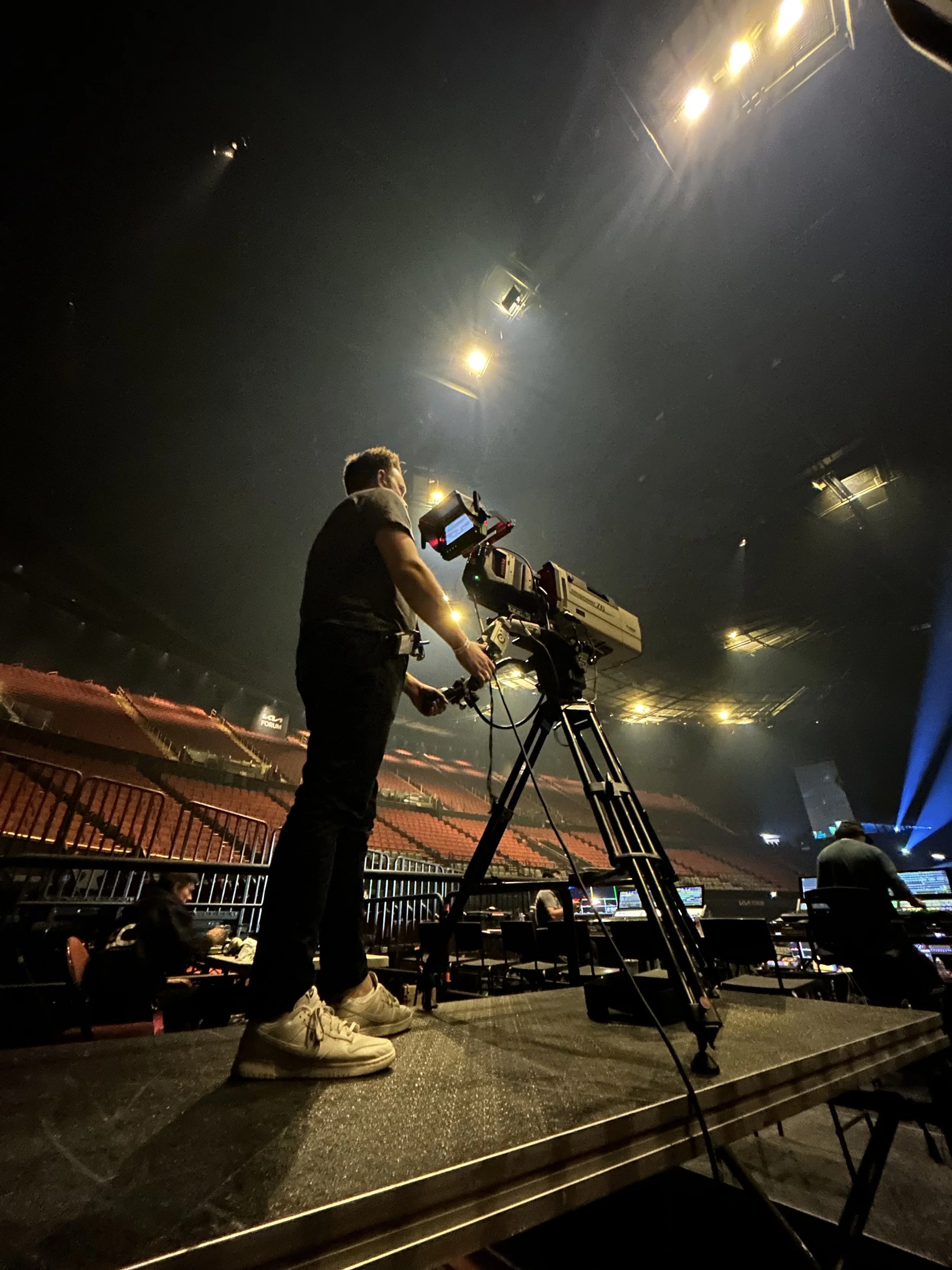 A person operating camera equipment on a stage in an indoor arena with empty red seating and stage lights.