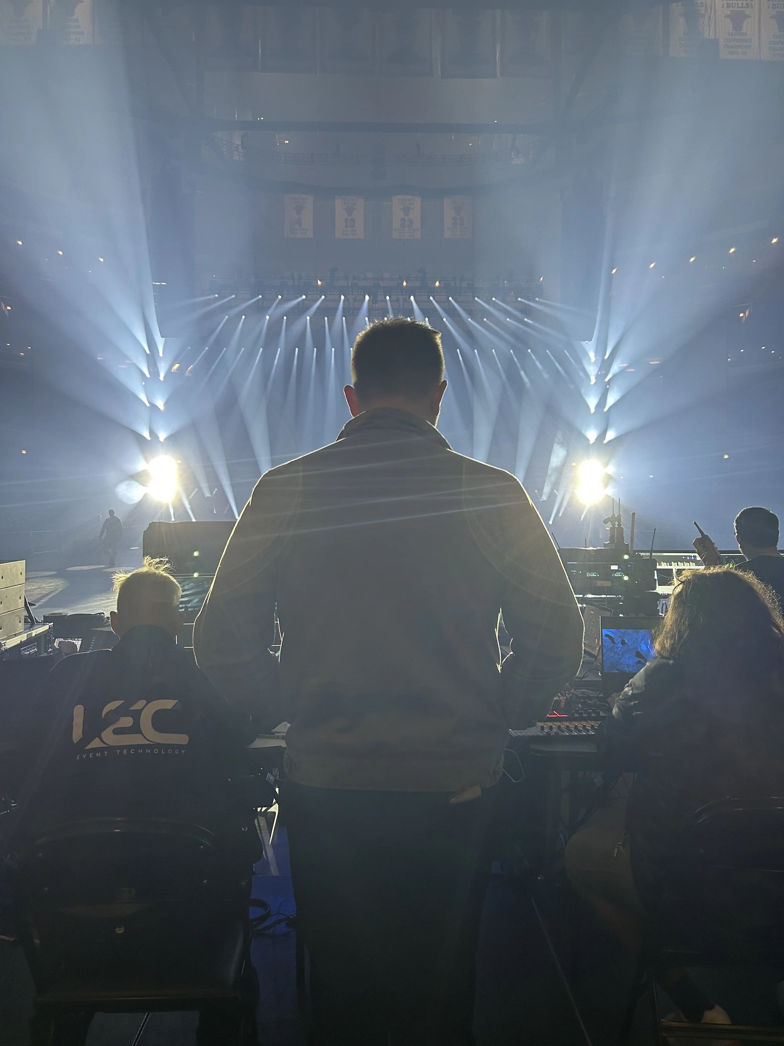 A person in a hoodie operating DJ equipment in front of bright stage lights at an indoor concert or event.